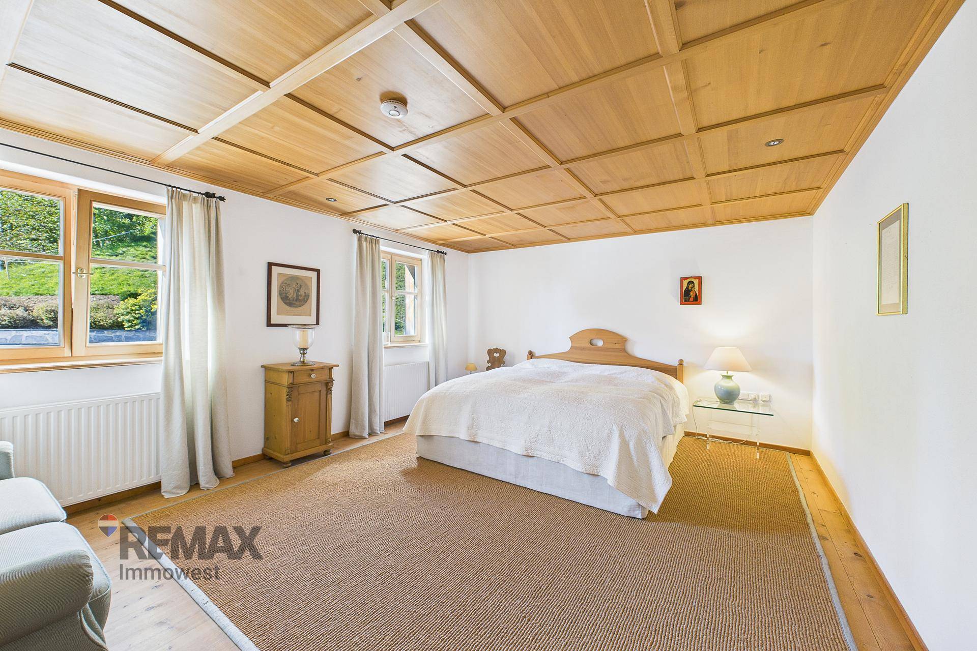 Geräumiges Schlafzimmer mit Holzkassettendecke, weiß gestrichenen Wänden und Blick ins Grüne.