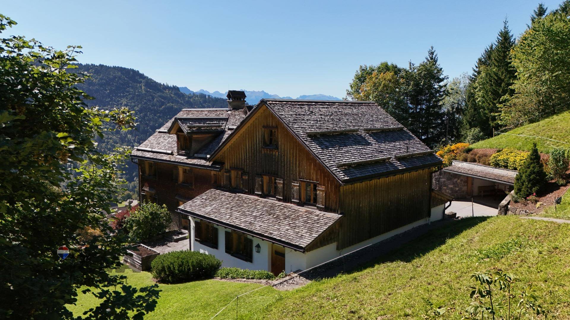 Rustikales Holzhaus mit traditioneller Architektur, umgeben von grünen Bäumen und einem Carport.