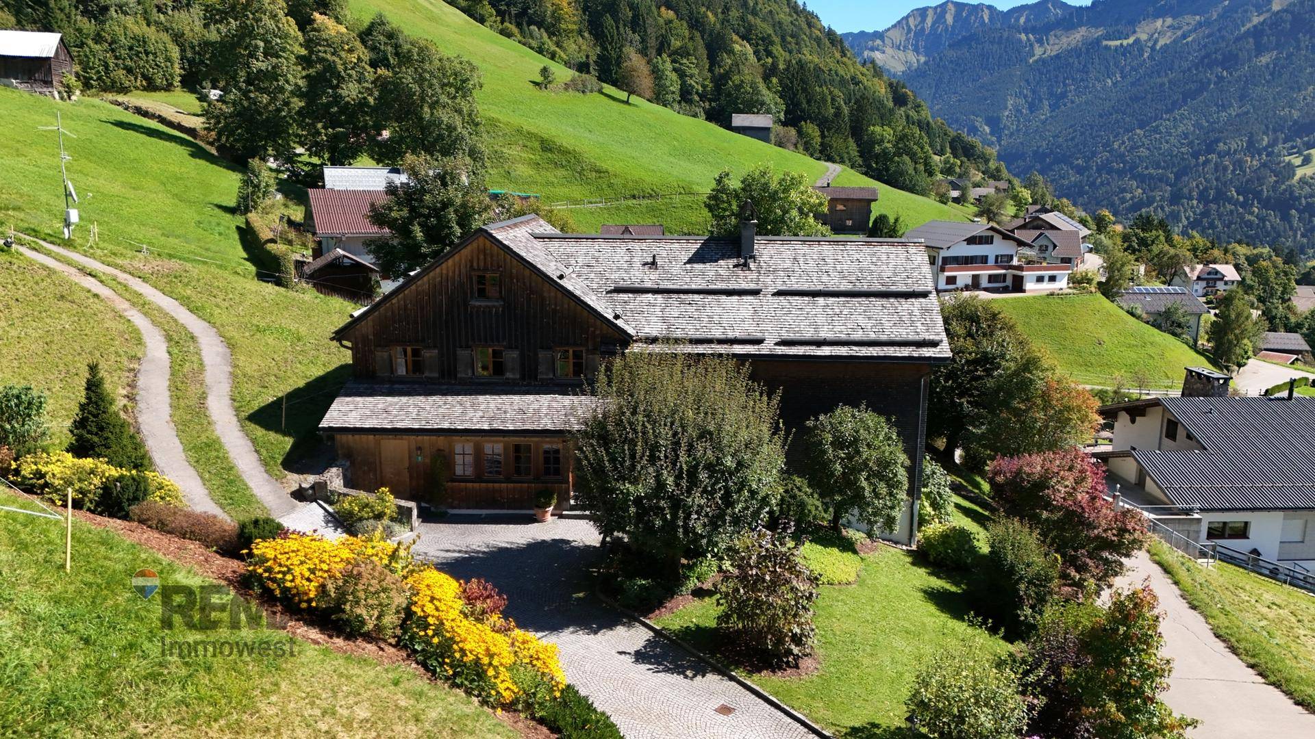 Anwesen mit traditionellem Holzhaus, weitläufigem Garten und gepflasterter Auffahrt in idyllischer Berglandschaft.
