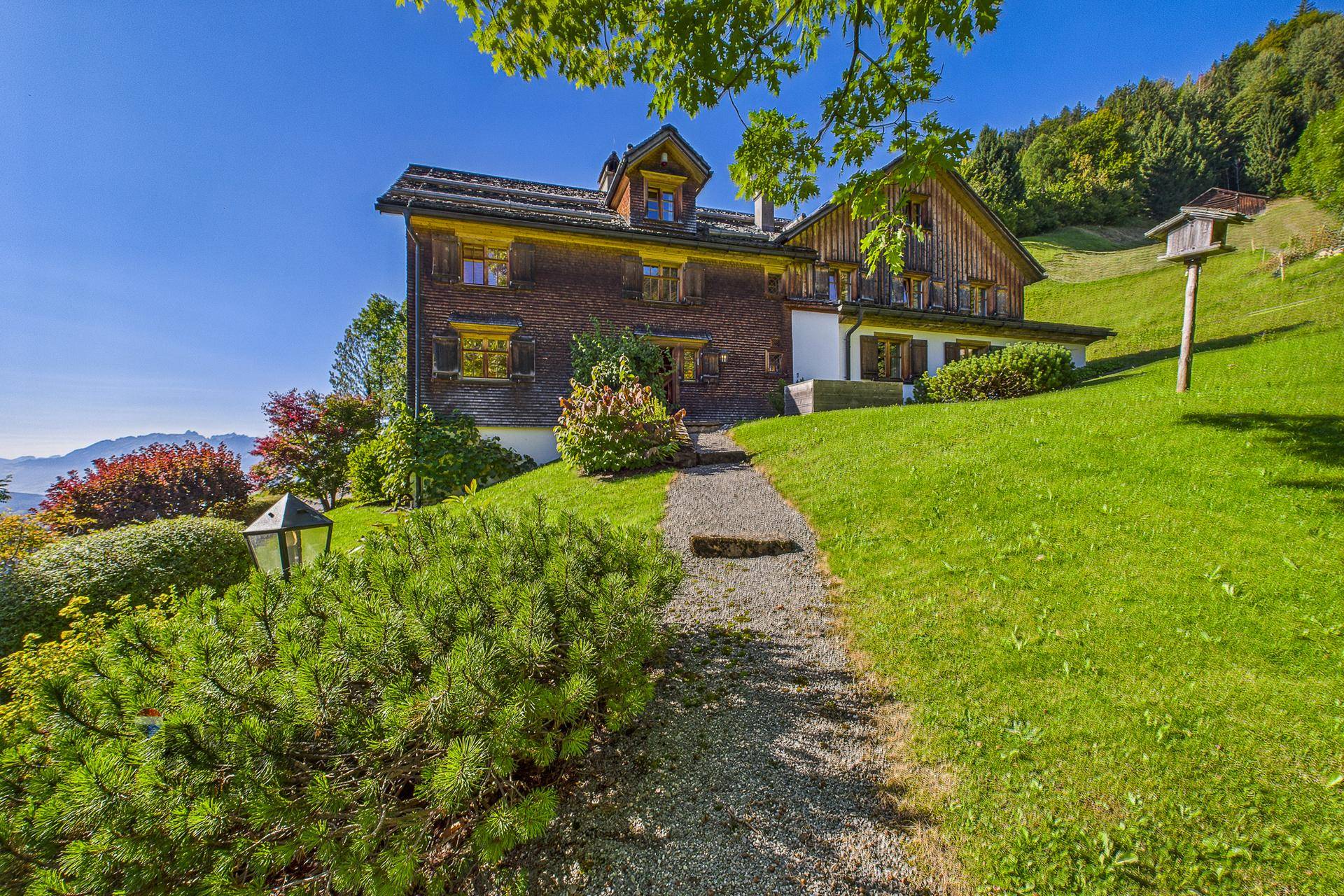 Idyllischer Gartenweg führt zum charmanten Holzhaus, umgeben von üppigem Grün und Bergblick.