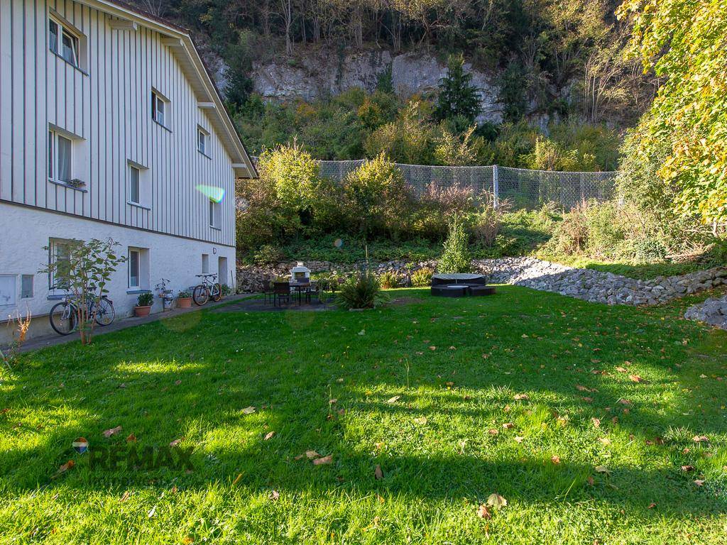 Sonniger Gartenbereich hinter dem Haus mit Rasen, Sitzgelegenheit und Blick auf die bewaldeten Hänge.