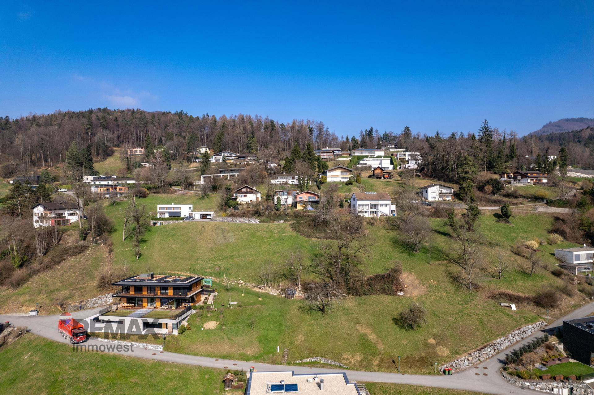 Panoramablick auf eine Wohnsiedlung am Hang mit Wald im Hintergrund und klarem blauen Himmel.