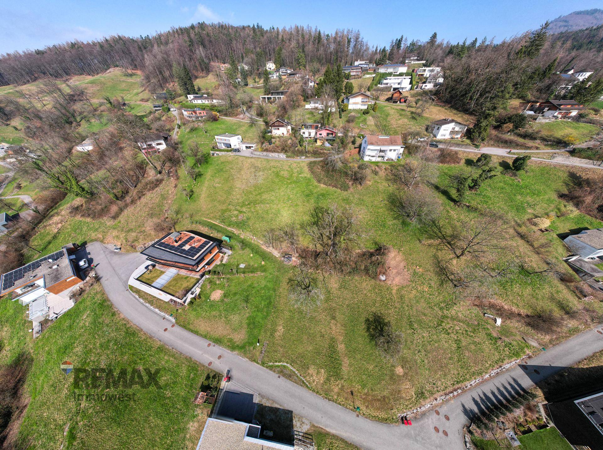 Luftaufnahme einer Wohngegend in Hanglage mit Häusern, grünen Grundstücken und Zufahrtsstraßen.