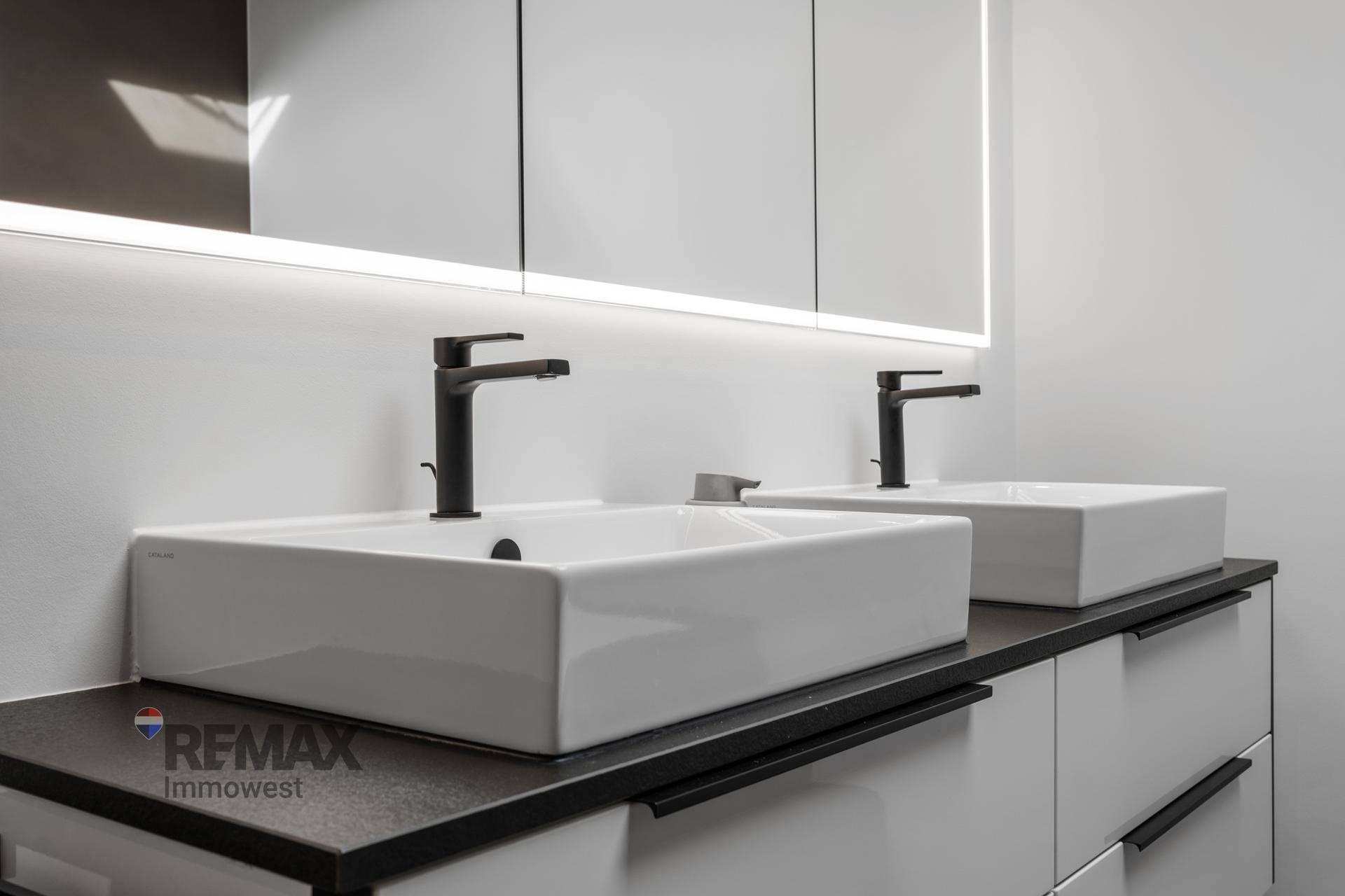 Modernes Badezimmer mit Doppelwaschbecken, schwarzen Armaturen und beleuchtetem Spiegelschrank.