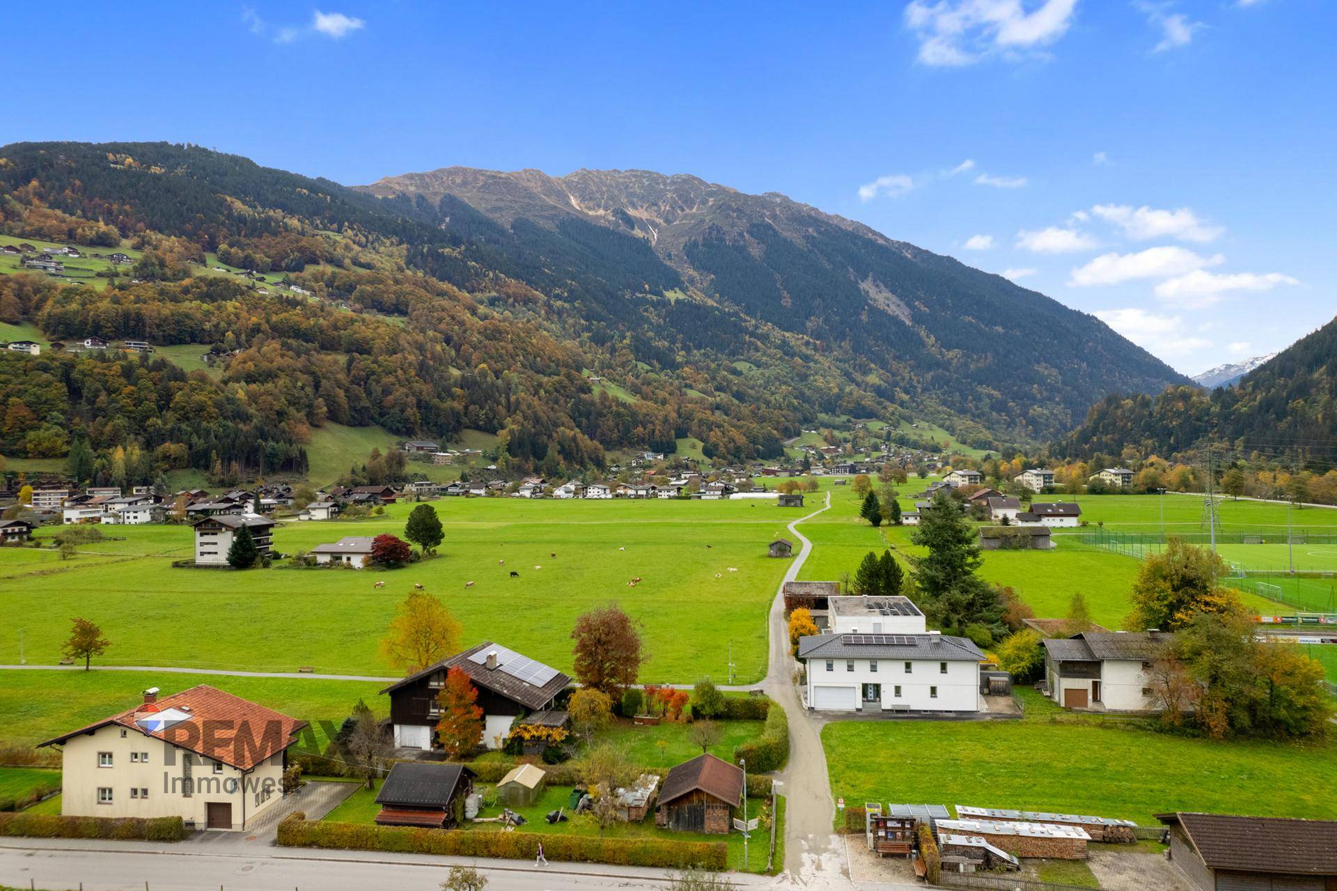 Weitläufige Luftaufnahme der ländlichen Umgebung mit grünen Feldern und verstreuten Häusern vor Bergkulisse.