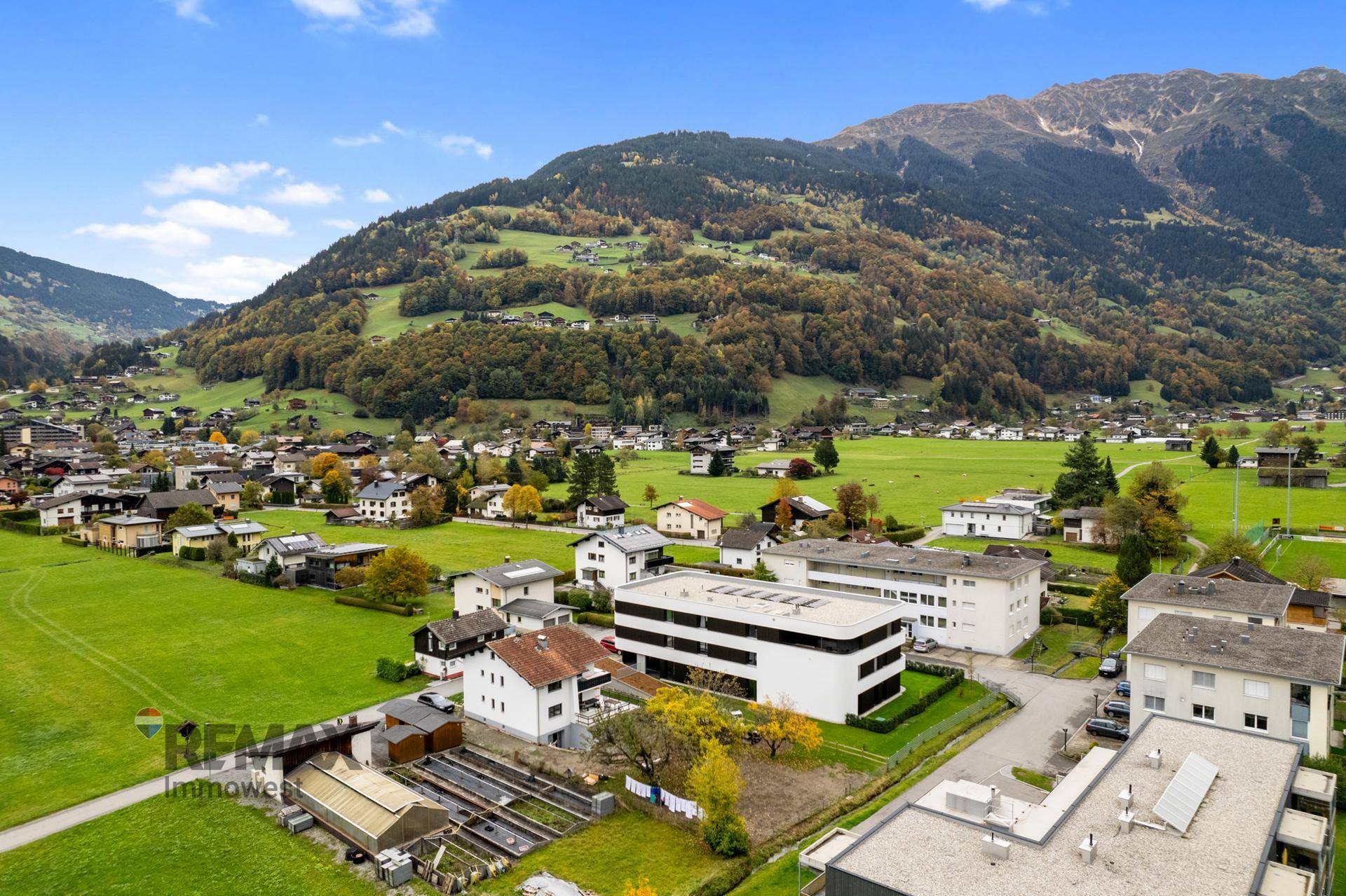 Luftaufnahme der Siedlung mit dem modernen Gebäude, umgeben von grünen Wiesen und Bergen.