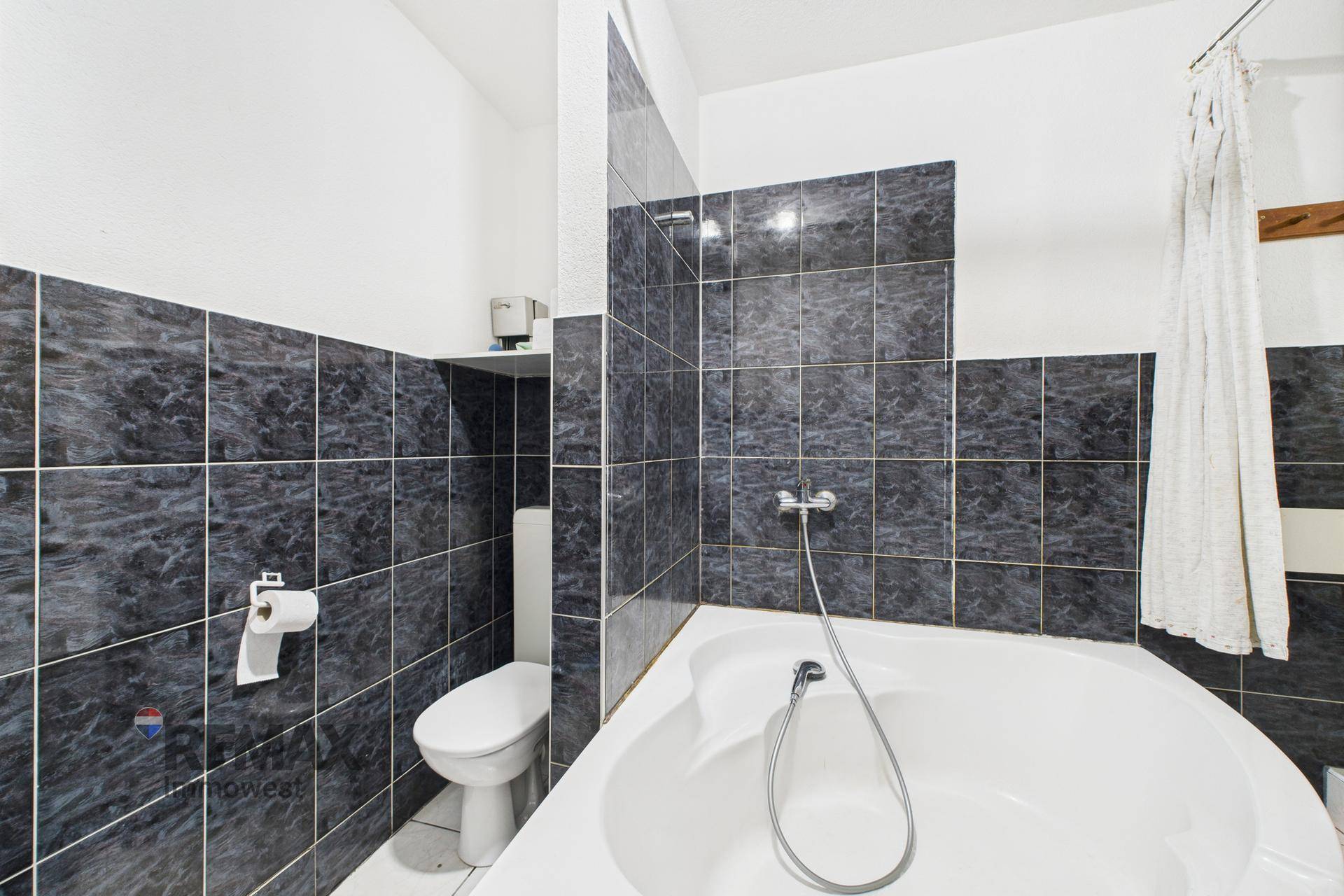 Badezimmer mit Eckbadewanne, dunklen Fliesen und Toilette, einfach und funktional.