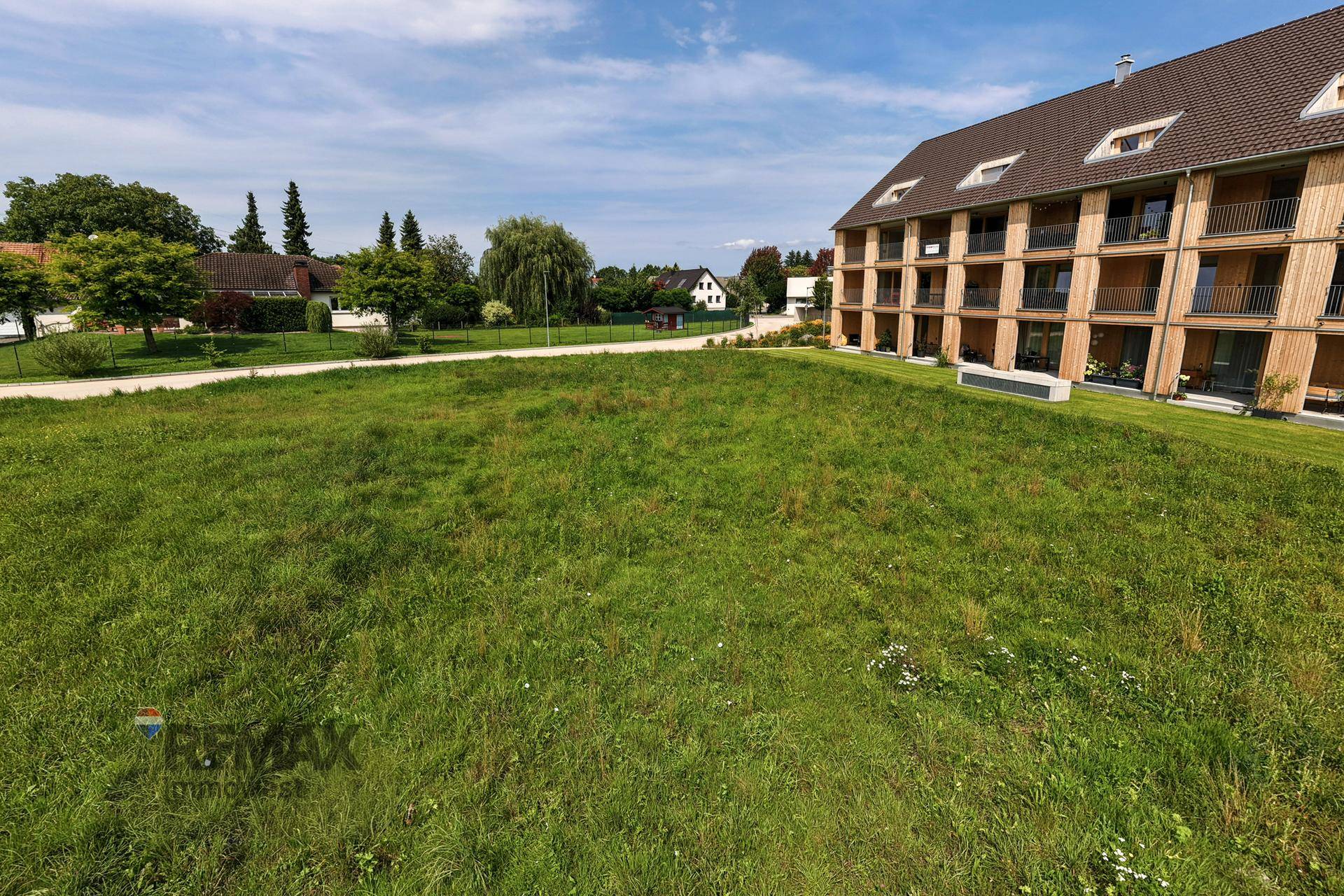 Grüne Wiese als Baugrundstück neben einem modernen Mehrfamilienhaus mit Holzfassade.