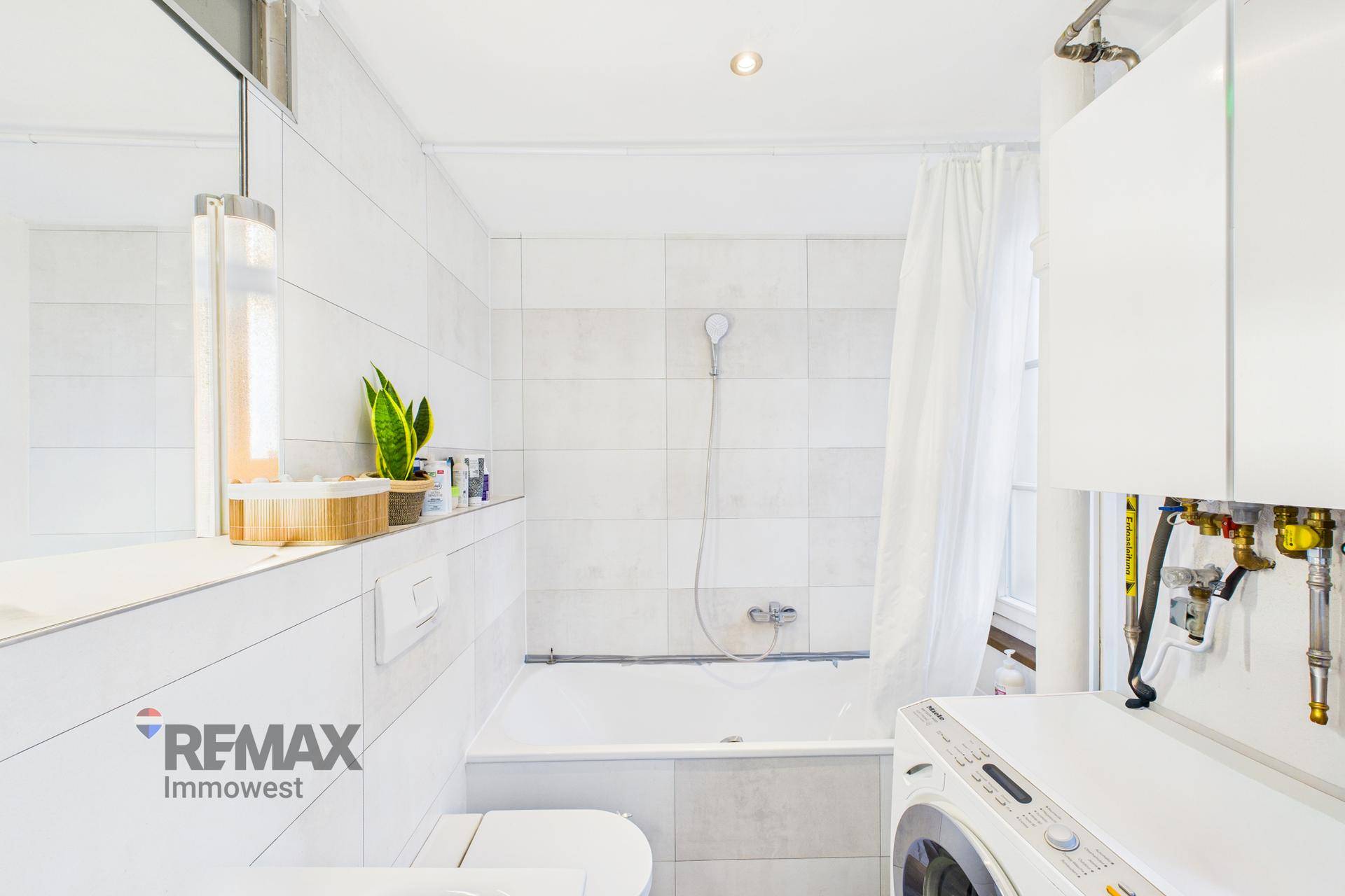 Helles Badezimmer mit Badewanne, Dusche und Waschmaschine in weiß gefliestem Raum.