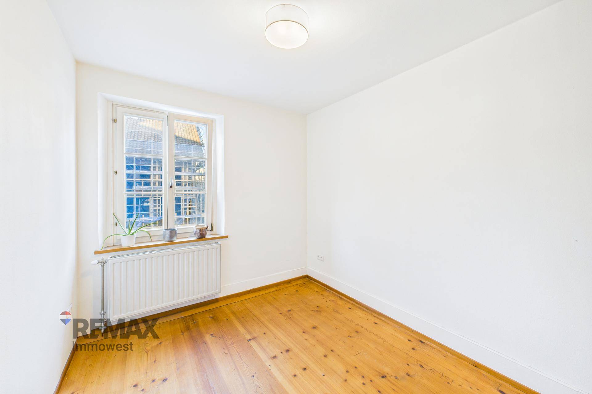 Leeres, helles Zimmer mit Holzfußboden und einem Fenster mit Heizkörper darunter.