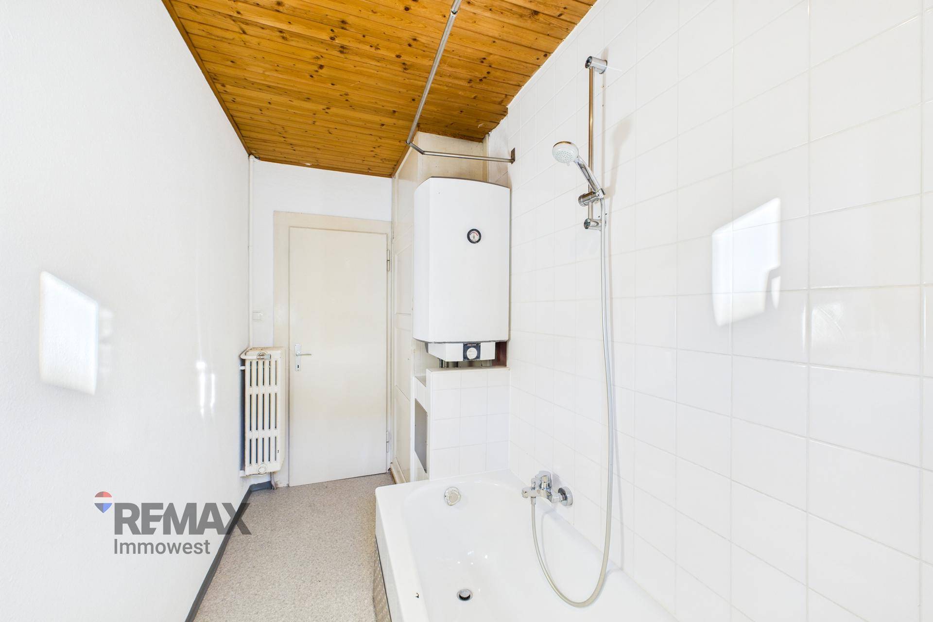 Badezimmer mit Badewanne, Duscharmatur und einem Warmwasserboiler an der Wand.