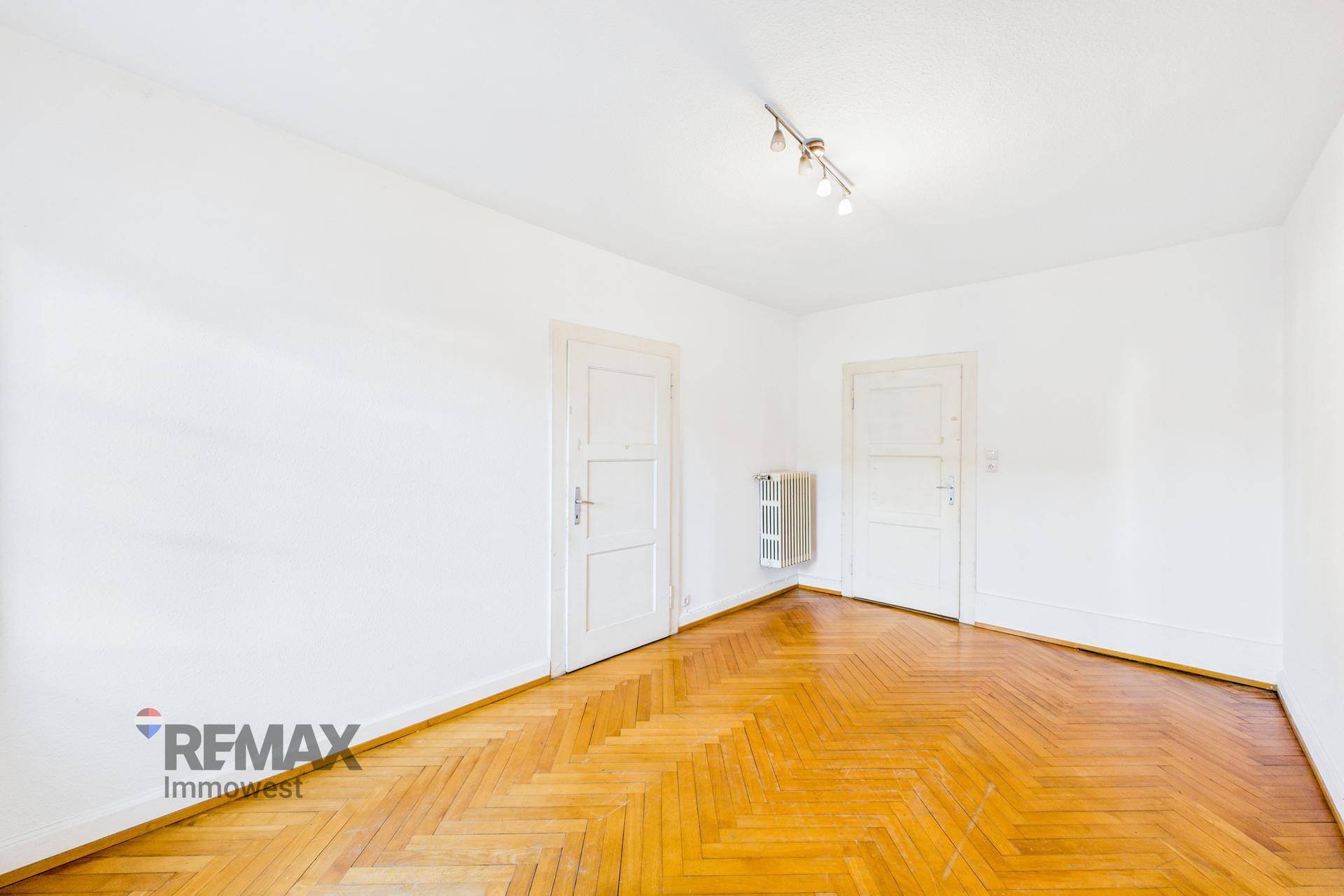 Geräumiges Zimmer mit Fischgrätparkettboden, weißen Wänden und zwei Türen.