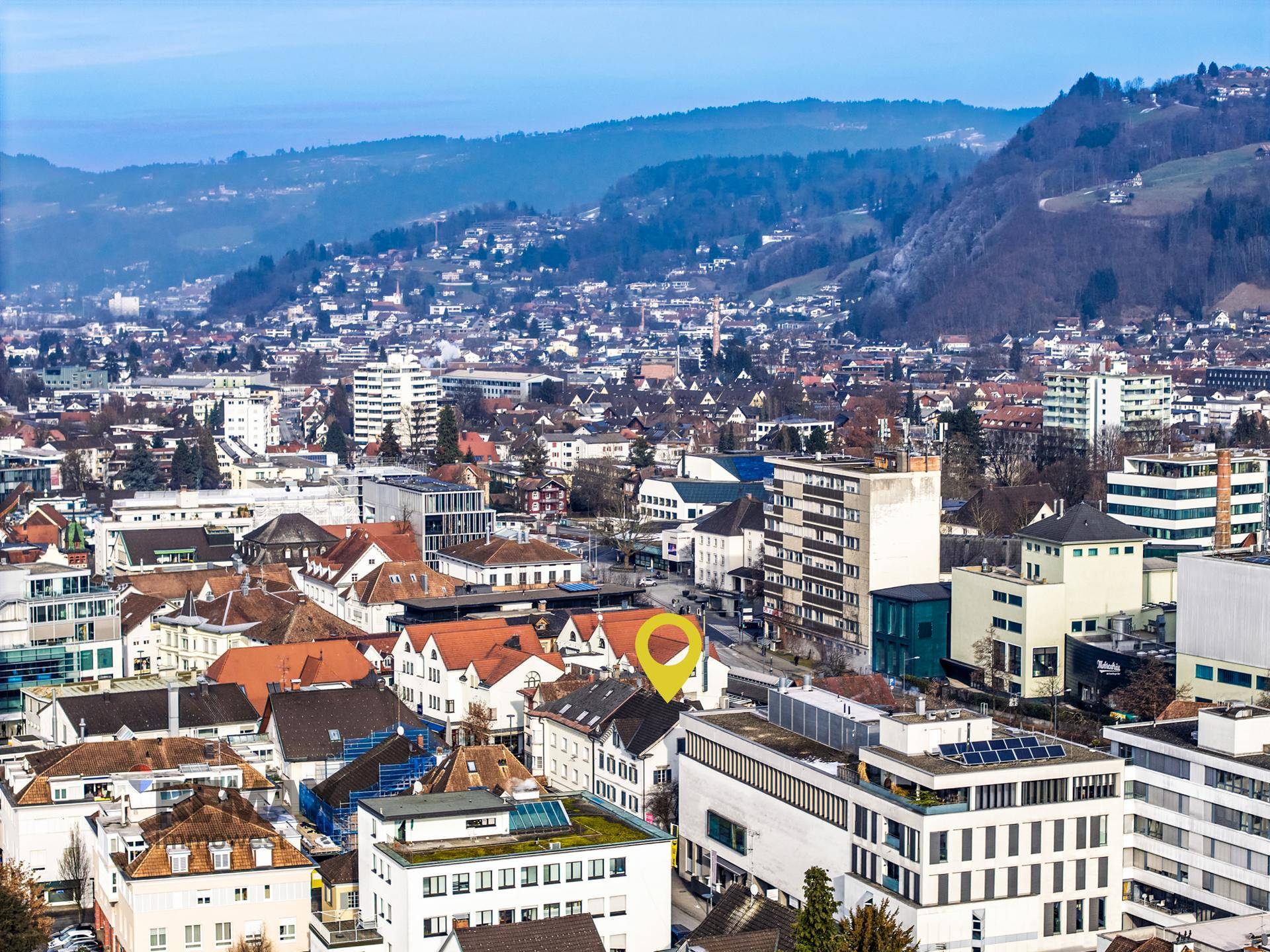 Panoramablick auf eine Stadtlandschaft mit Bergen im Hintergrund und markiertem Standort.