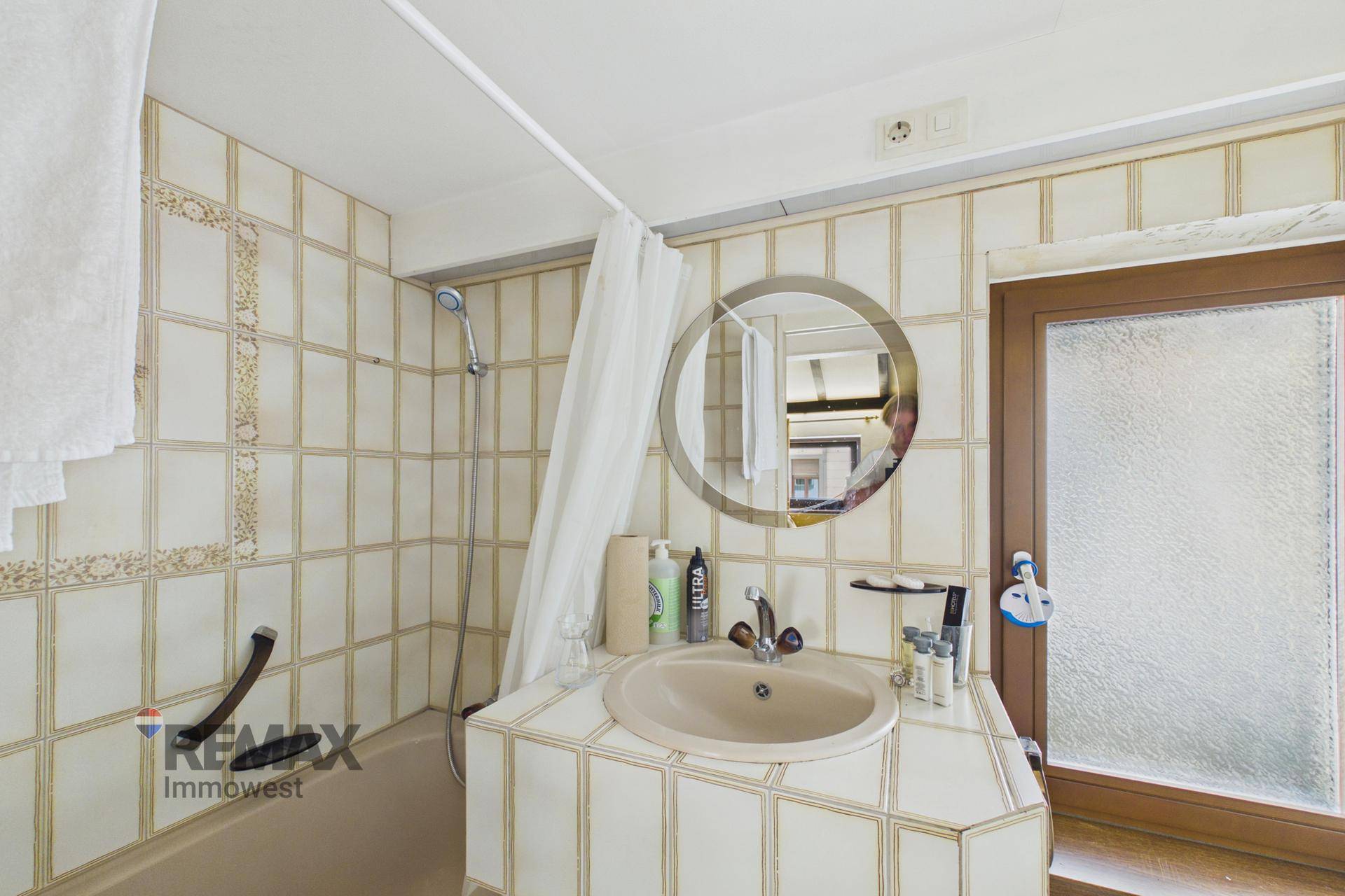 Badezimmer mit beigefarbenen Fliesen, Badewanne mit Duschfunktion und Fenster.