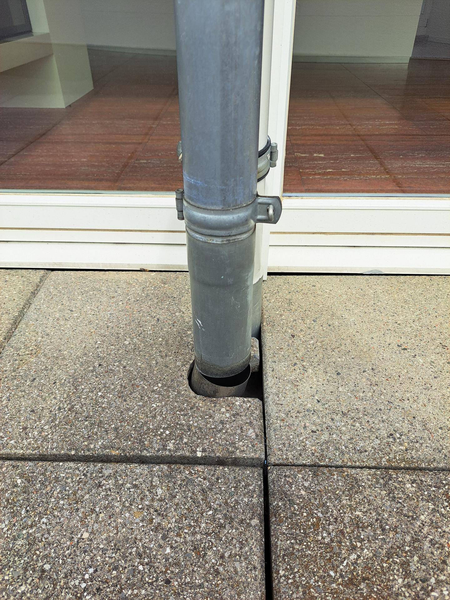 Regenfallrohr an der Fassade mit Betonplatten im Vordergrund und reflektiertem Holzfußboden im Inneren.