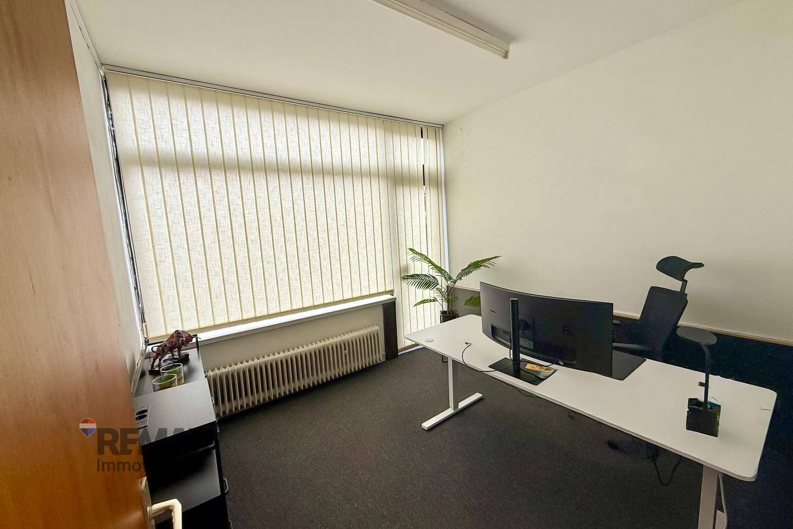 Modern eingerichtetes Büro mit Teppichboden, Jalousien und ergonomischem Arbeitsplatz mit zwei Monitoren.