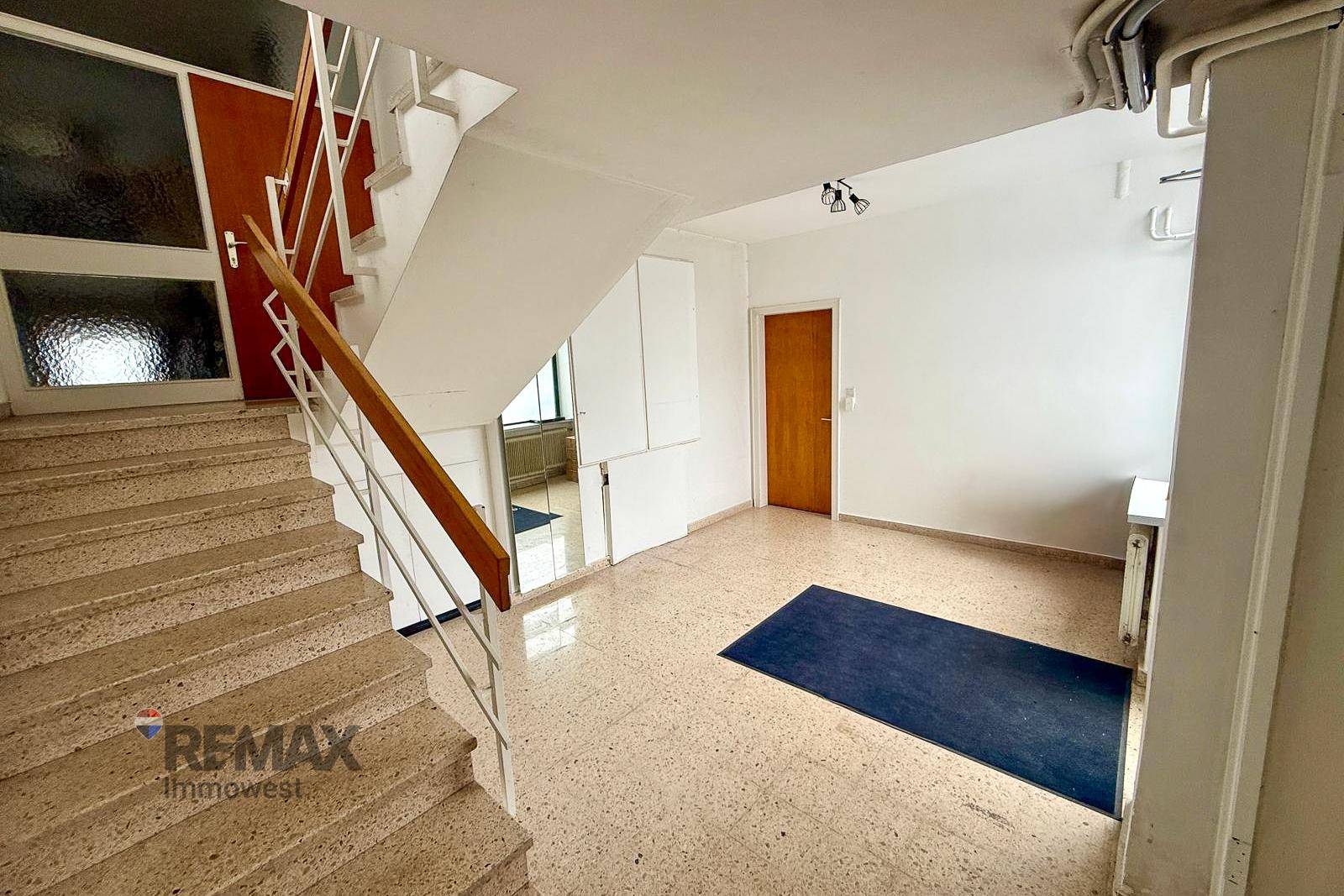 Treppenhaus und Flur mit Terrazzoboden und Holzgeländer in einem Mehrfamilienhaus.
