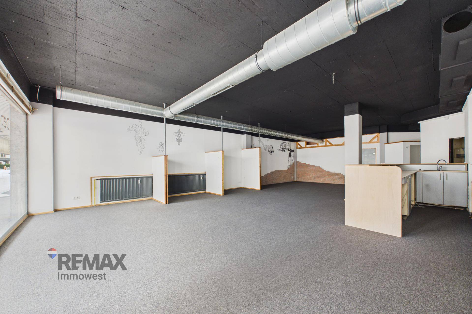Geräumiger Geschäftsraum mit grauem Teppichboden, schwarzer Decke und sichtbaren Lüftungsrohren, inklusive kleiner Küchenzeile.