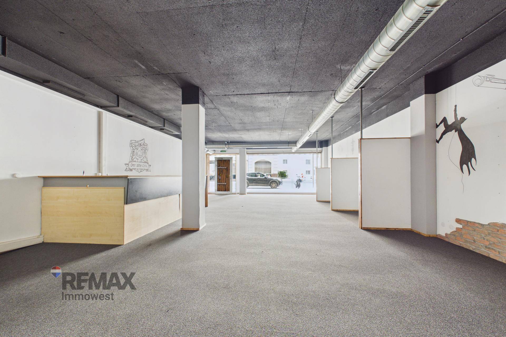 Weitläufiger Geschäftsraum mit grauem Teppich, schwarzer Decke und industriellen Lüftungsrohren, gut beleuchtet.