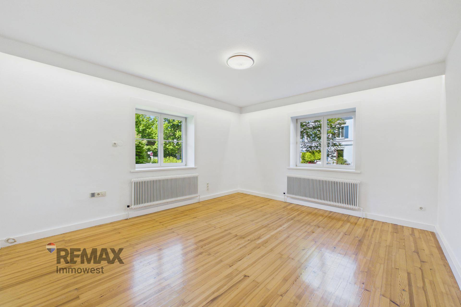 Geräumiges Wohnzimmer mit zwei großen Fenstern, die viel Tageslicht hereinlassen, und Holzfußboden.