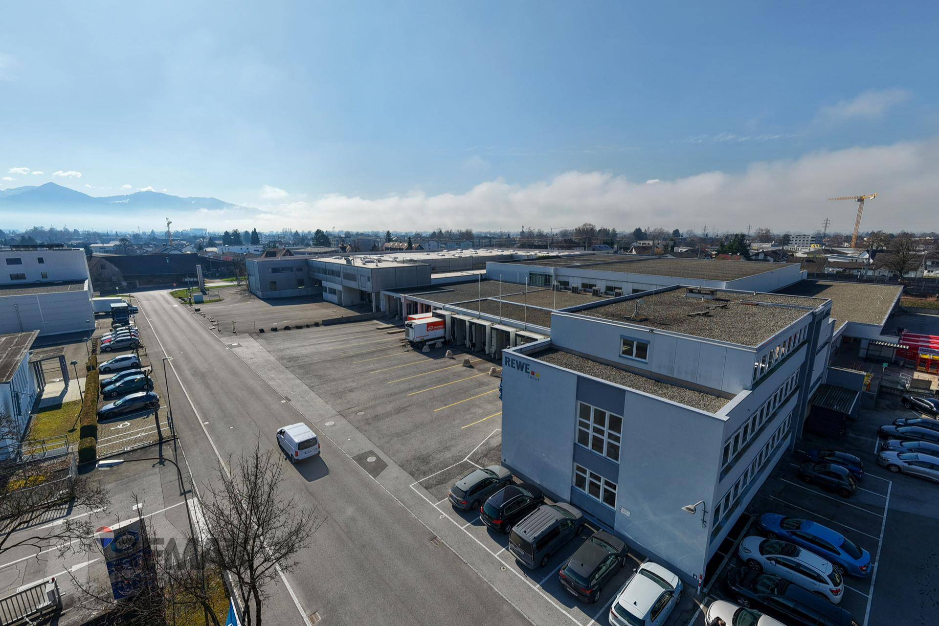 Weitläufiges Gewerbegebiet mit mehreren Gebäuden, Parkplätzen und Bergen im Hintergrund.