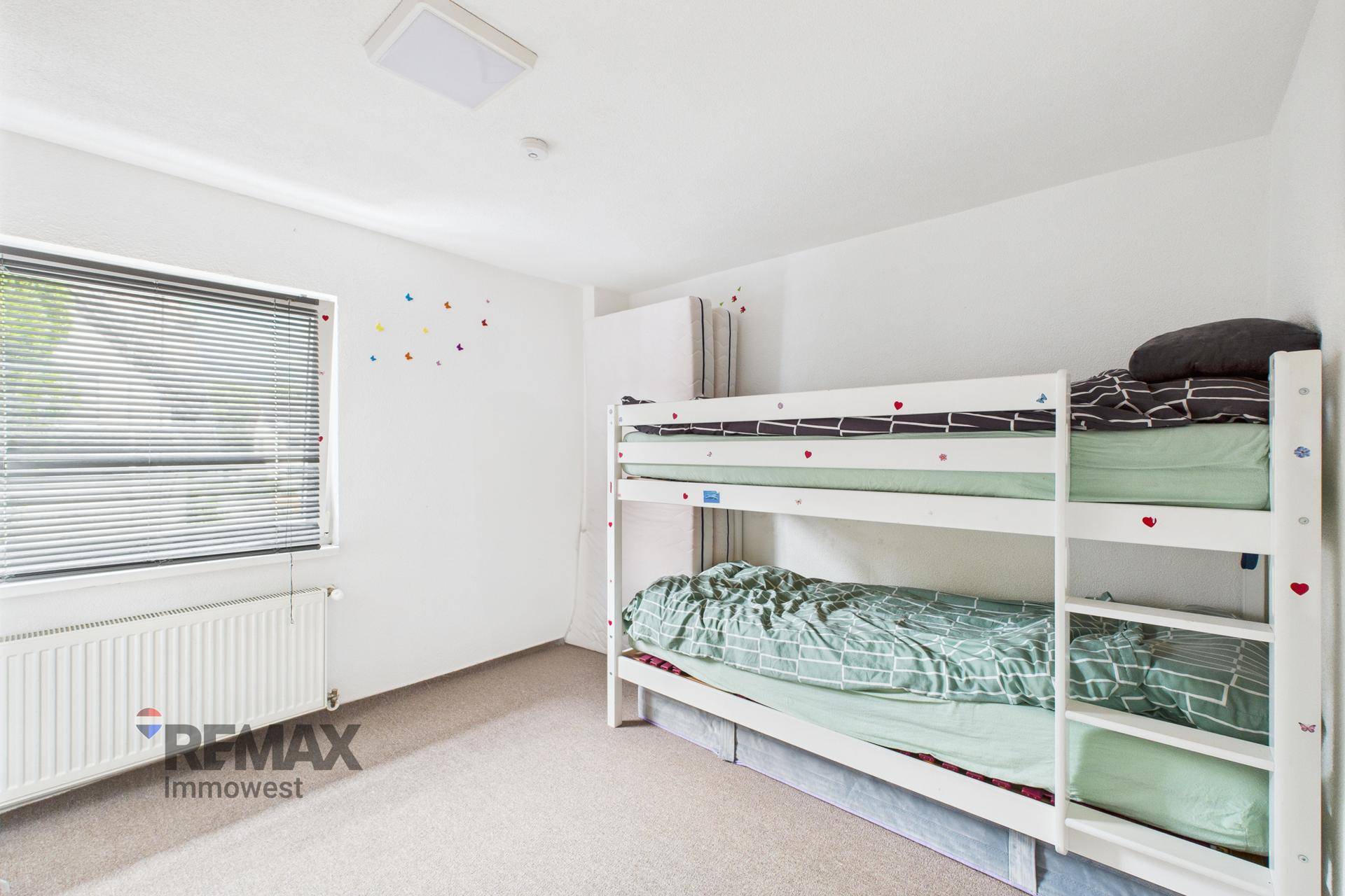Kinderzimmer mit Etagenbett, Teppichboden und einem Fenster mit Jalousien.