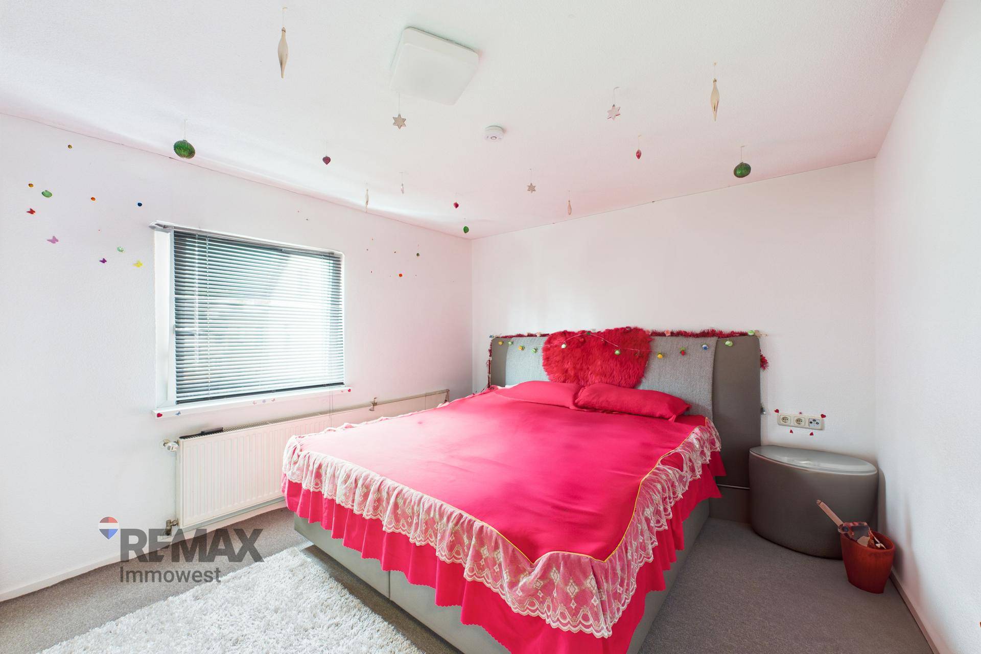 Schlafzimmer mit Doppelbett, rosa Bettwäsche, Teppichboden und einem Fenster mit Jalousien.