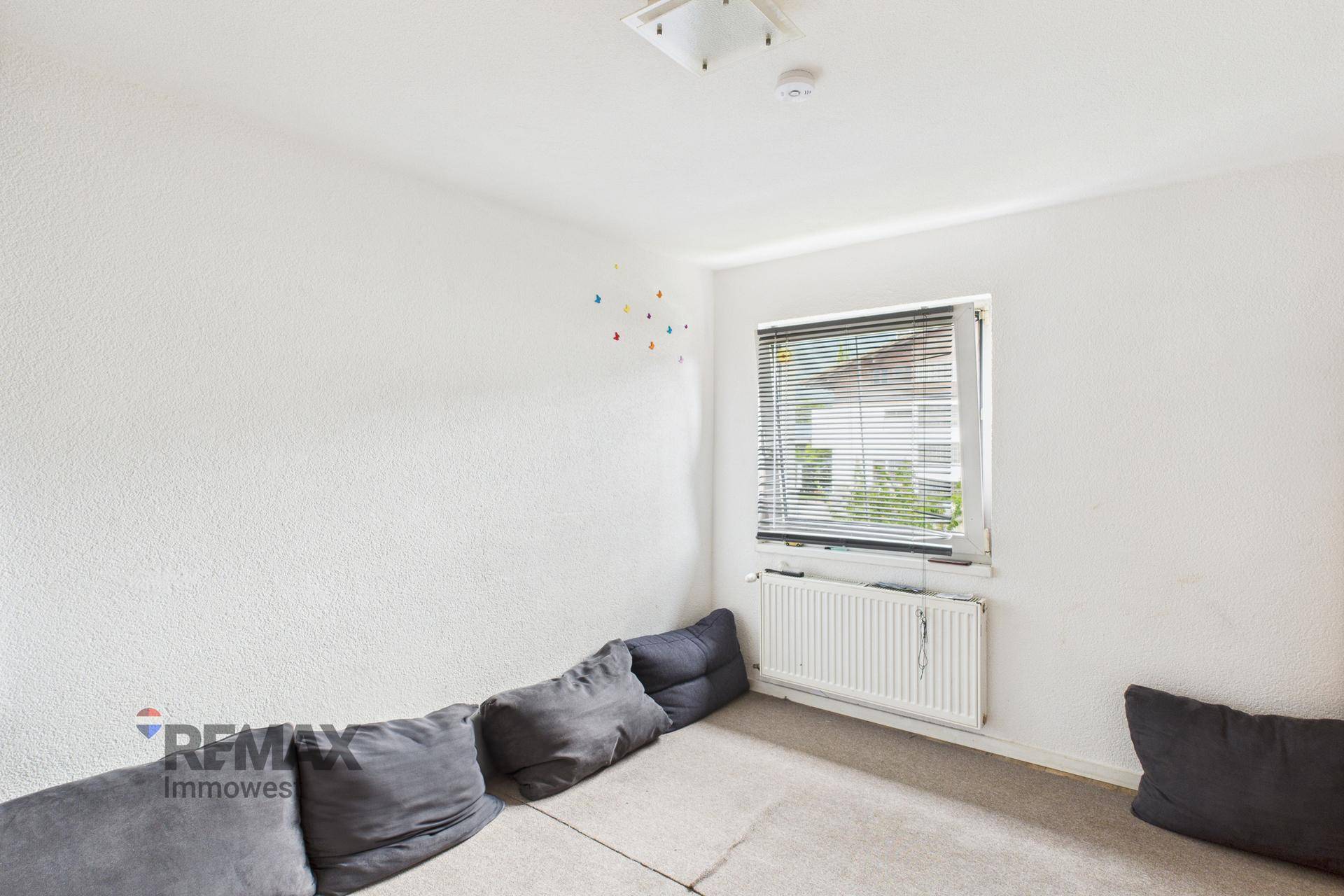 Schlichtes Zimmer mit Teppichboden, weiß gestrichenen Wänden und einem Fenster mit Jalousien.
