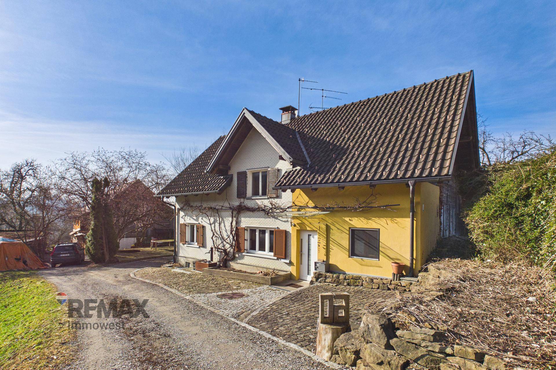 Vorderansicht des Hauses mit gelber Fassade und gepflastertem Vorplatz unter blauem Himmel.