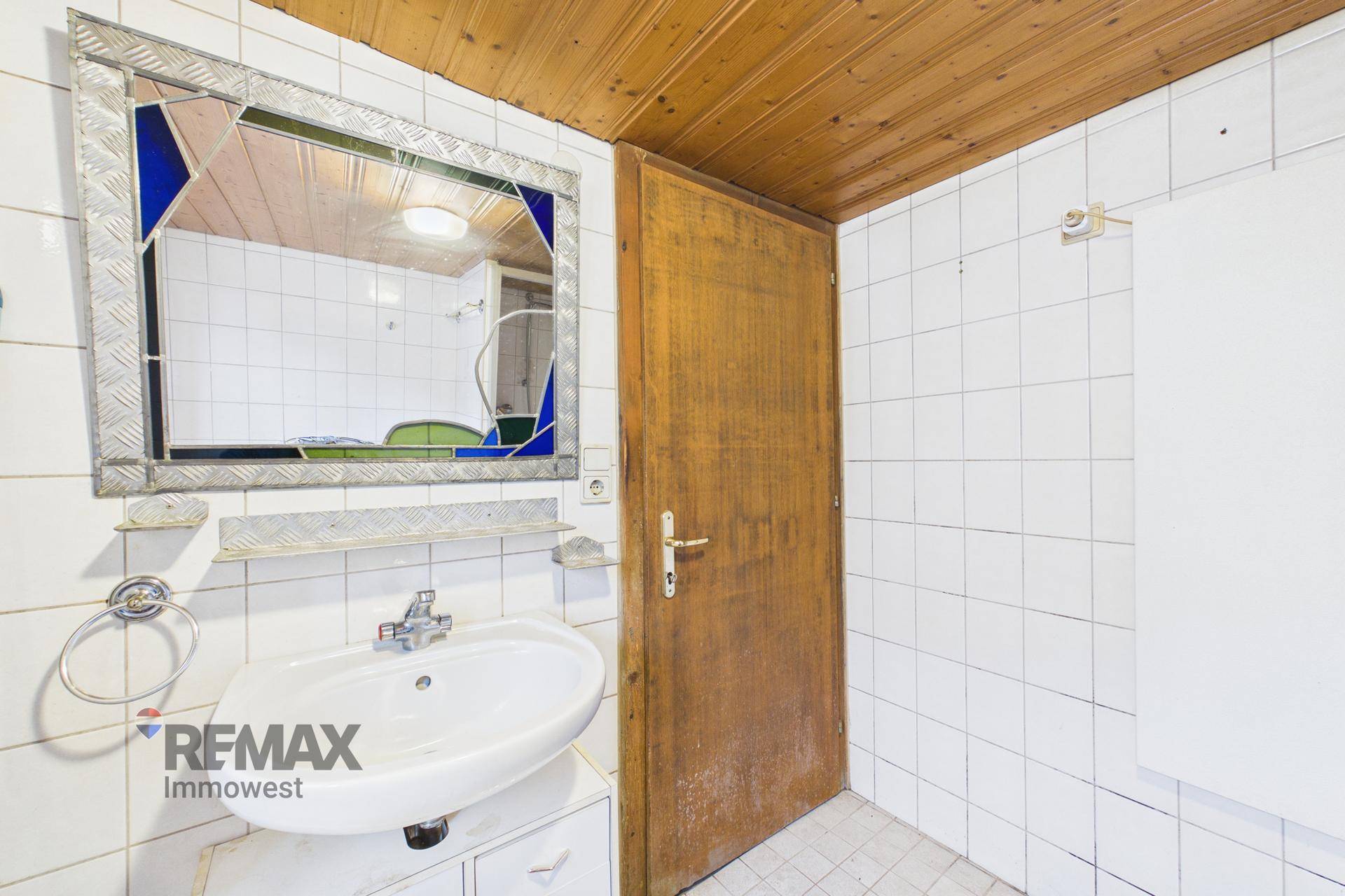 Badezimmer mit Waschbecken, Spiegel und Holzdecke, gefliest in Weiß.