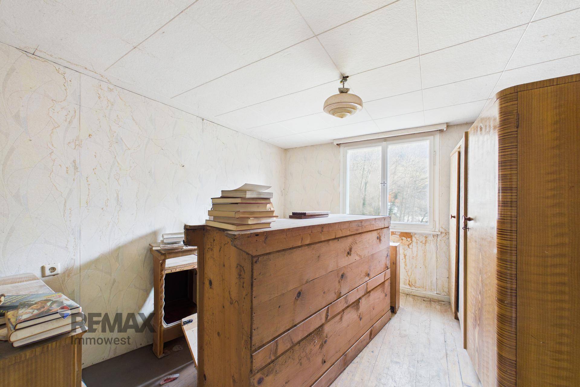 Schlafzimmer mit abgenutzter Tapete, Holzmöbeln und einem Fenster, das auf die Umgebung blickt.