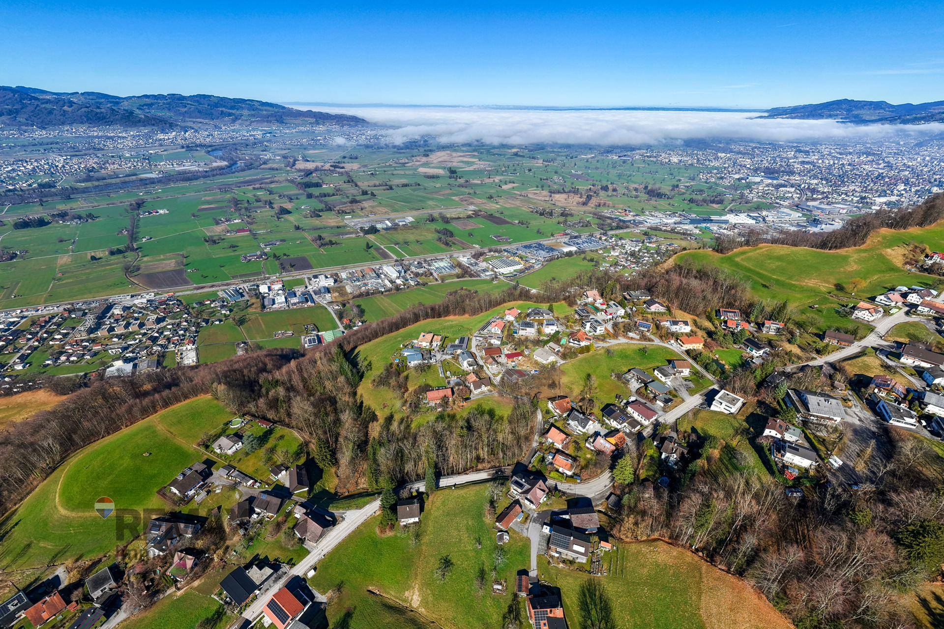Weitläufige Luftaufnahme einer Wohnsiedlung in einem grünen Tal mit Bergen im Hintergrund.