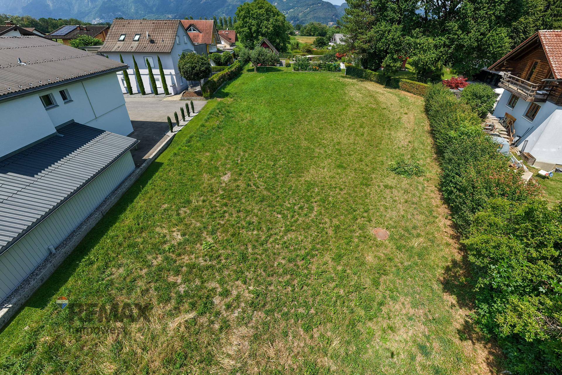 Großes Baugrundstück mit Rasenfläche, umgeben von Hecken und Bäumen, mit Bergblick.