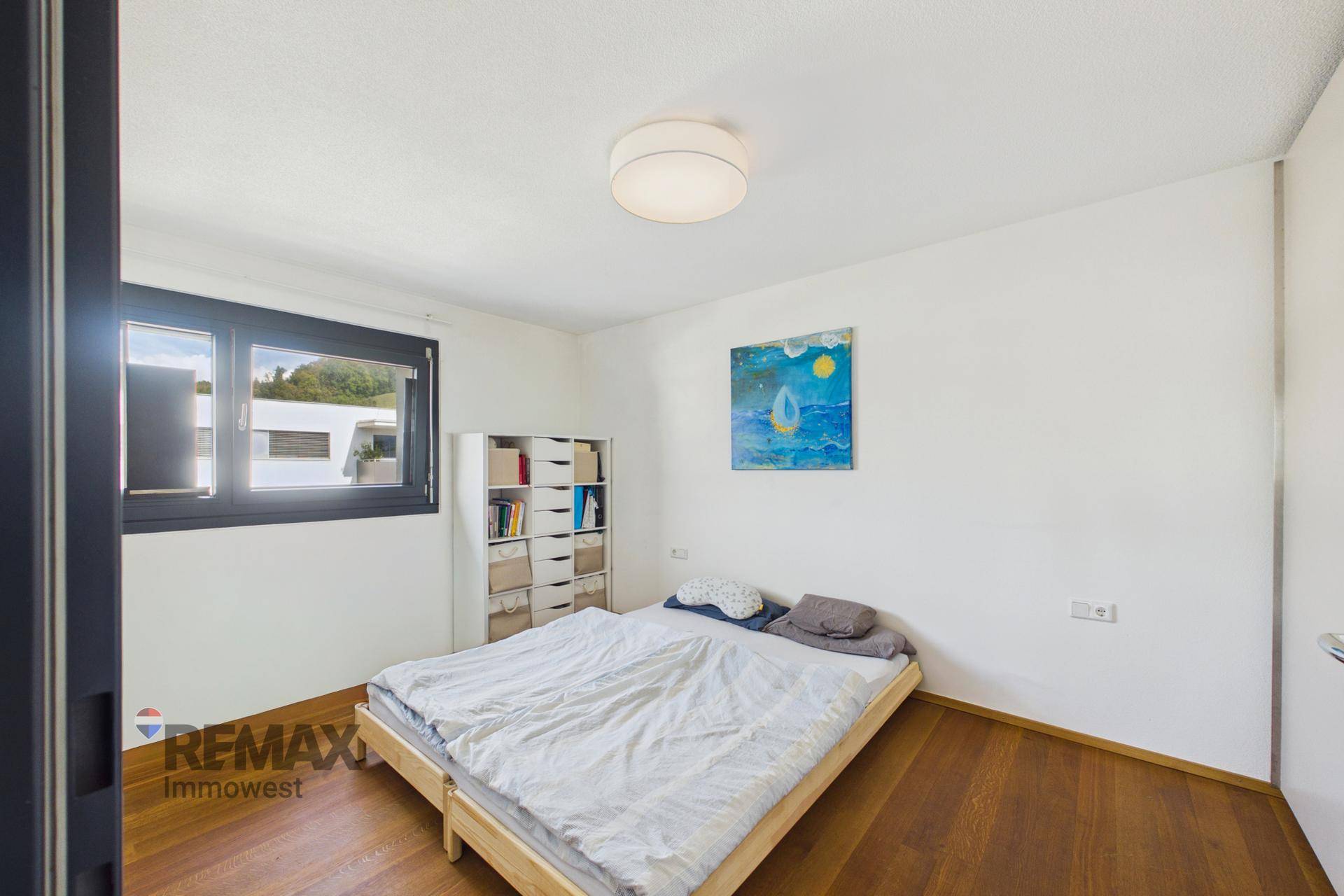 Helles Schlafzimmer mit Holzboden, Doppelbett und einem Fenster, das Tageslicht hereinlässt.