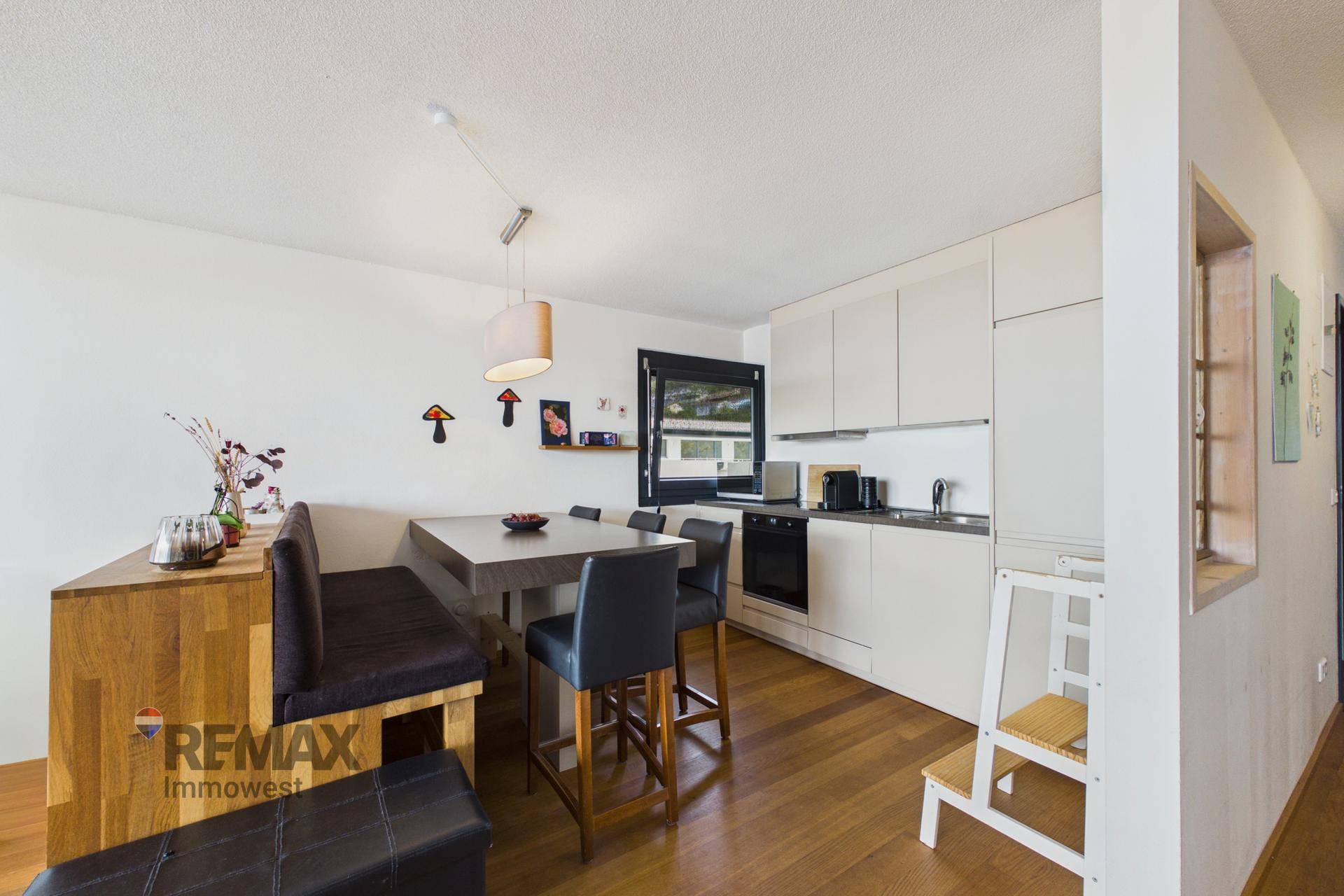 Moderne Küche mit weißer Einrichtung, Esstisch und Holzboden, gut beleuchtet durch ein Fenster.