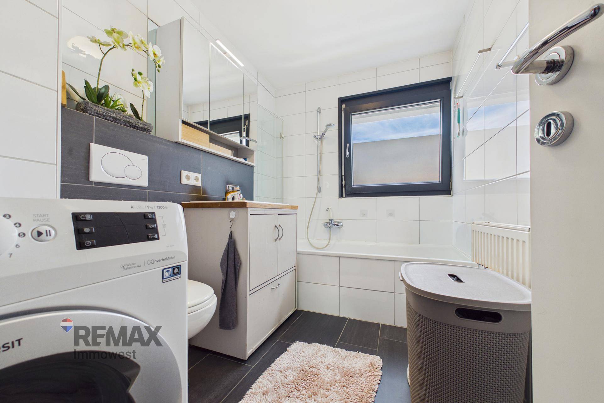 Modernes Badezimmer mit Badewanne, Waschmaschine und Fenster, hell gefliest.