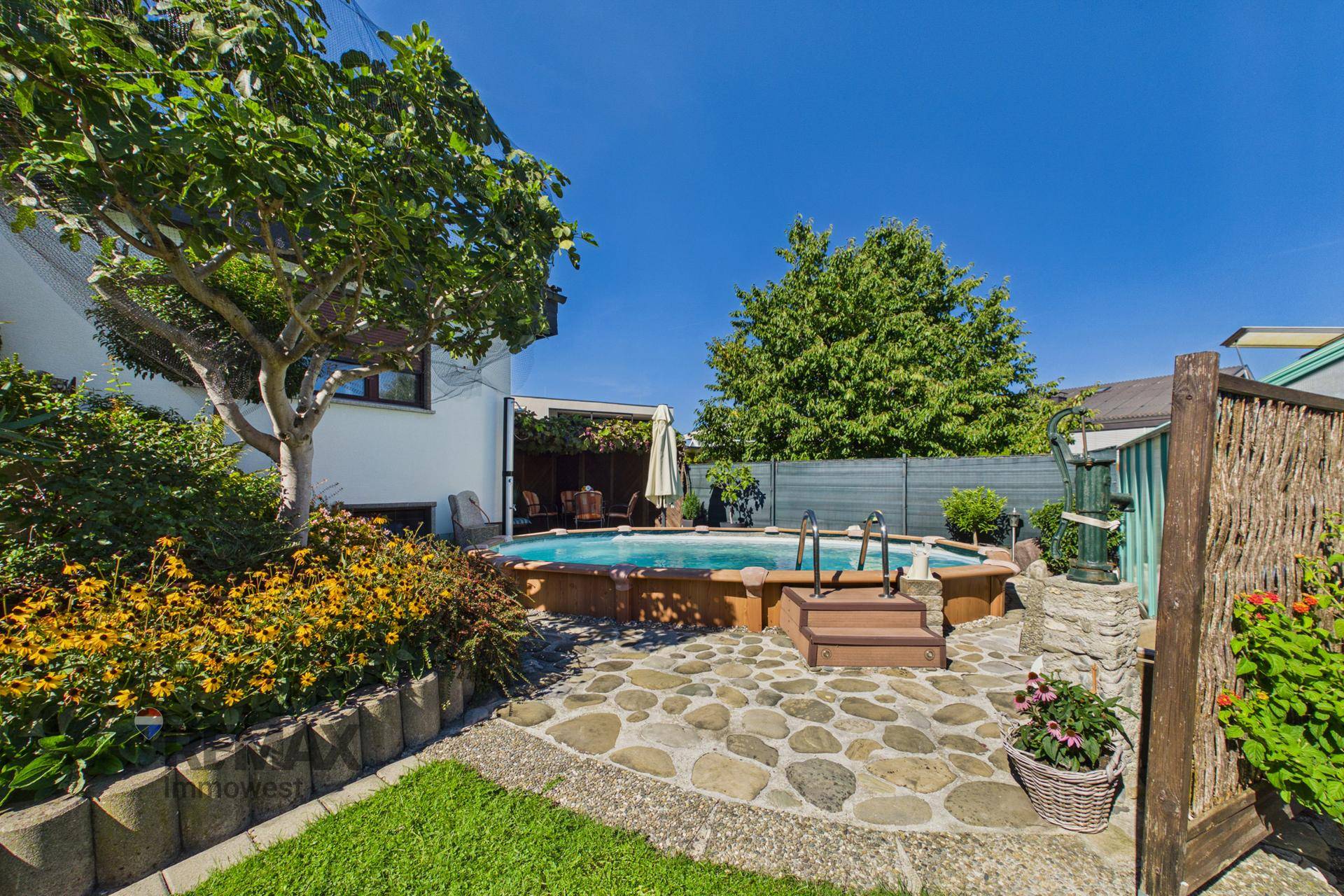 Idyllischer Gartenbereich mit oberirdischem Pool, Steinpflaster und üppiger Bepflanzung unter blauem Himmel.