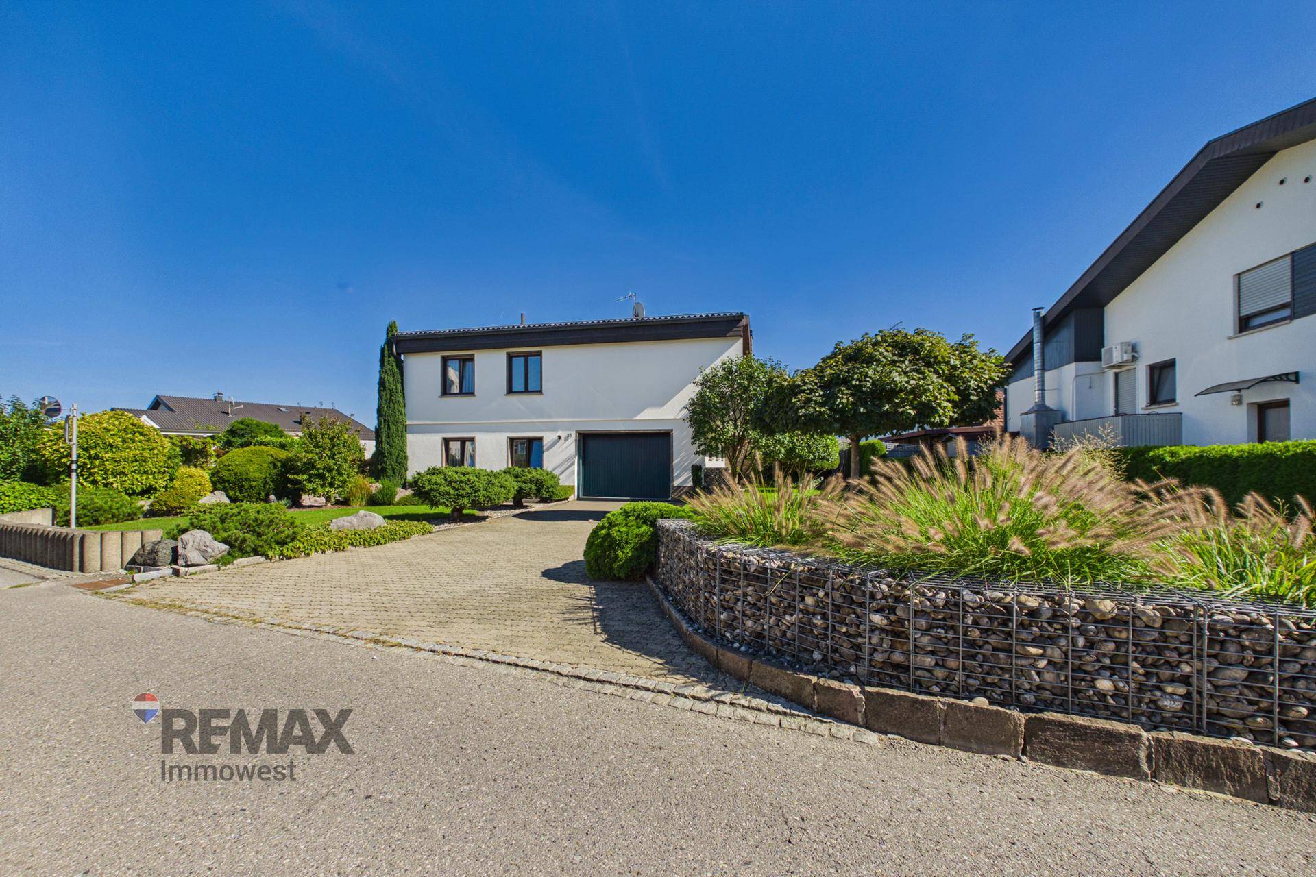 Einfamilienhaus mit gepflasterter Auffahrt, Garage und Gabionenmauer im Vorgarten an einem sonnigen Tag.