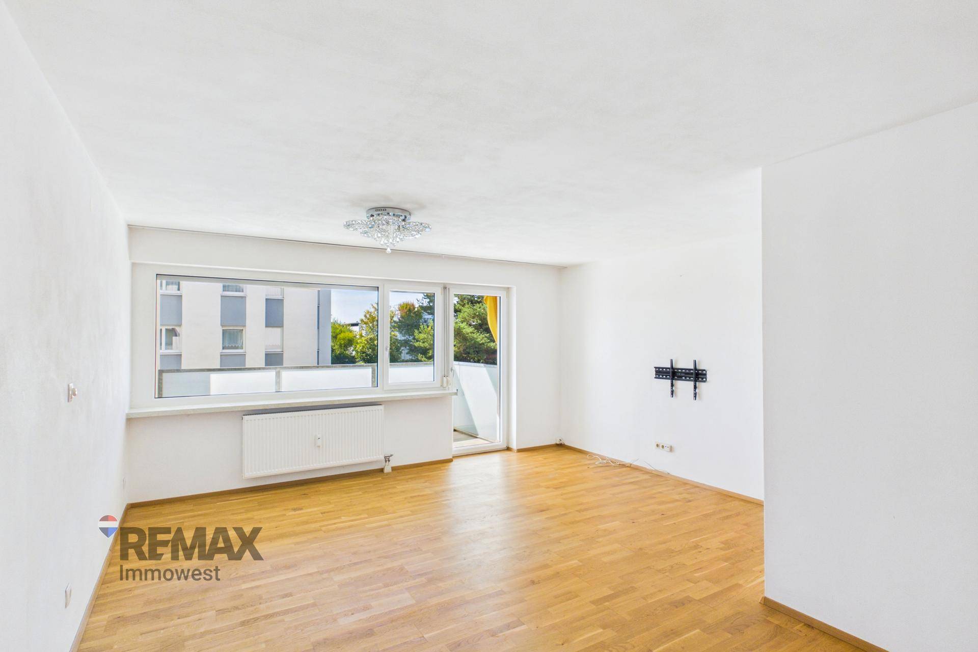 Geräumiges Wohnzimmer mit Parkettboden, weiß gestrichenen Wänden und Blick nach draußen.