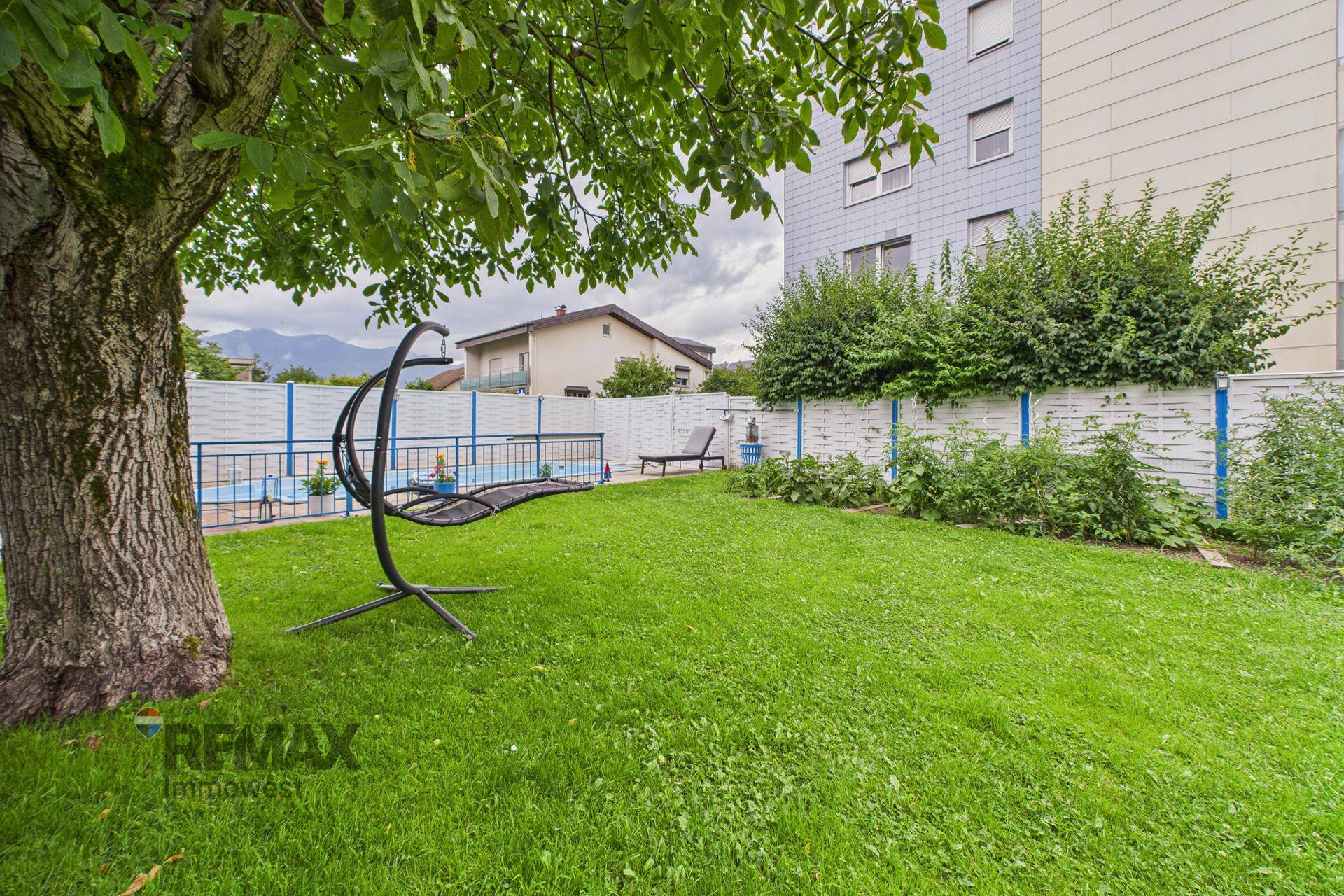 Gepflegter Garten mit Rasenfläche, großem Baum und Schaukelstuhl, umgeben von einem Zaun.