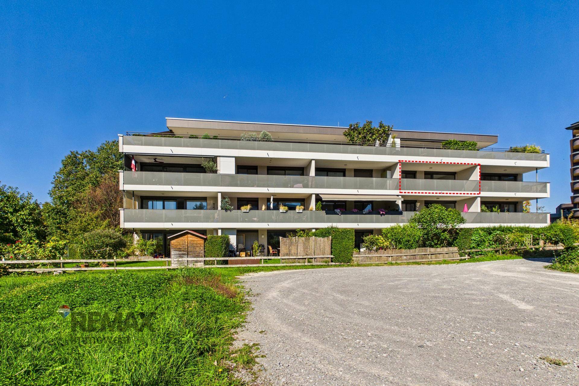 Modernes Mehrfamilienhaus mit mehreren Balkonen und grüner Umgebung unter blauem Himmel.