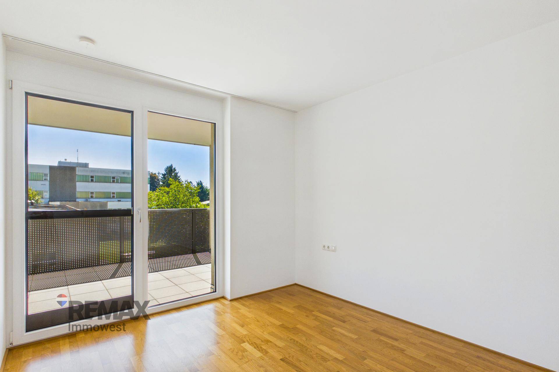 Helles Schlafzimmer mit Holzboden und großem Fenster, das Zugang zu einem Außenbereich ermöglicht.