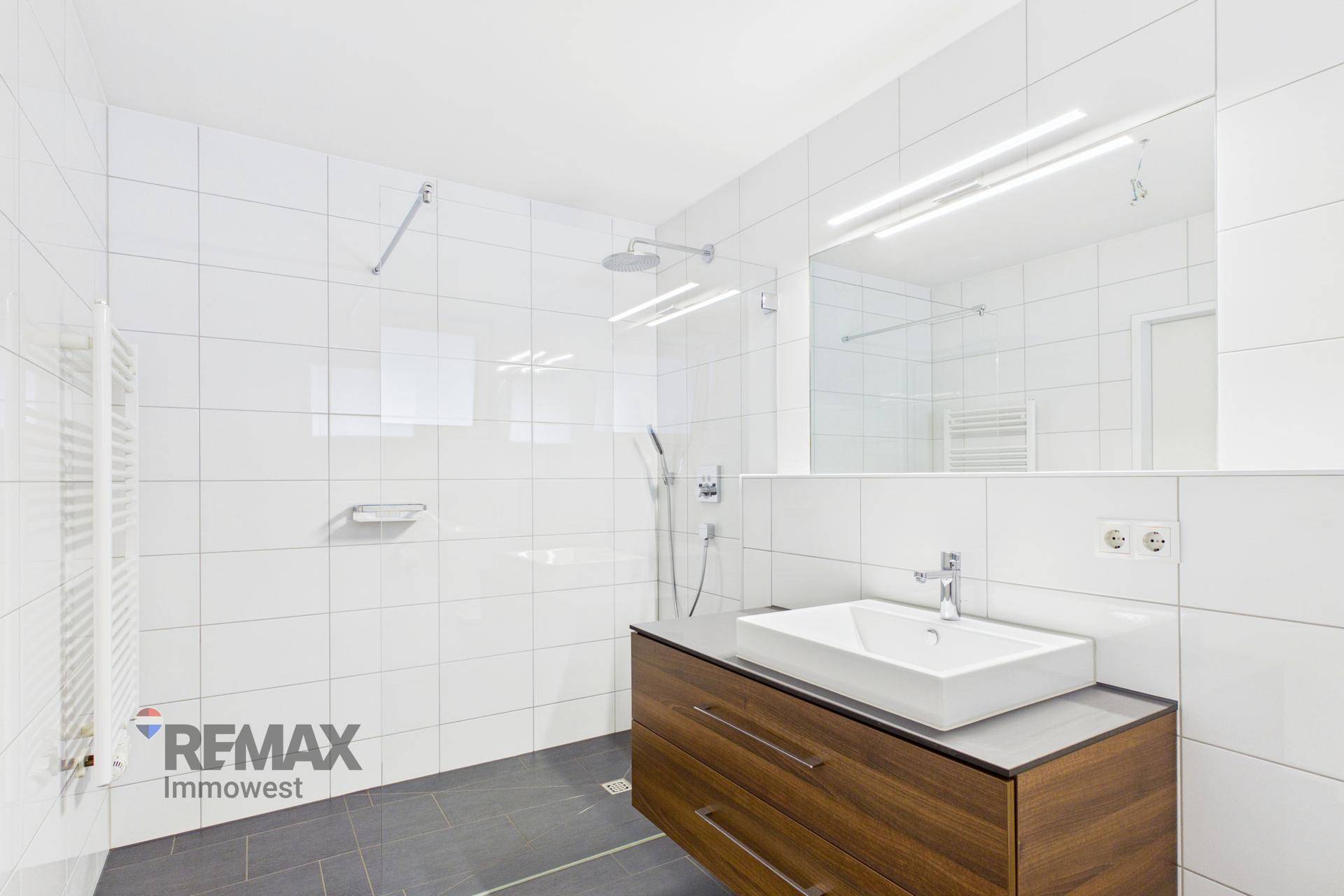 Modernes Badezimmer mit weißen Fliesen, dunklem Boden, Holzwaschtisch und einer geräumigen begehbaren Dusche.
