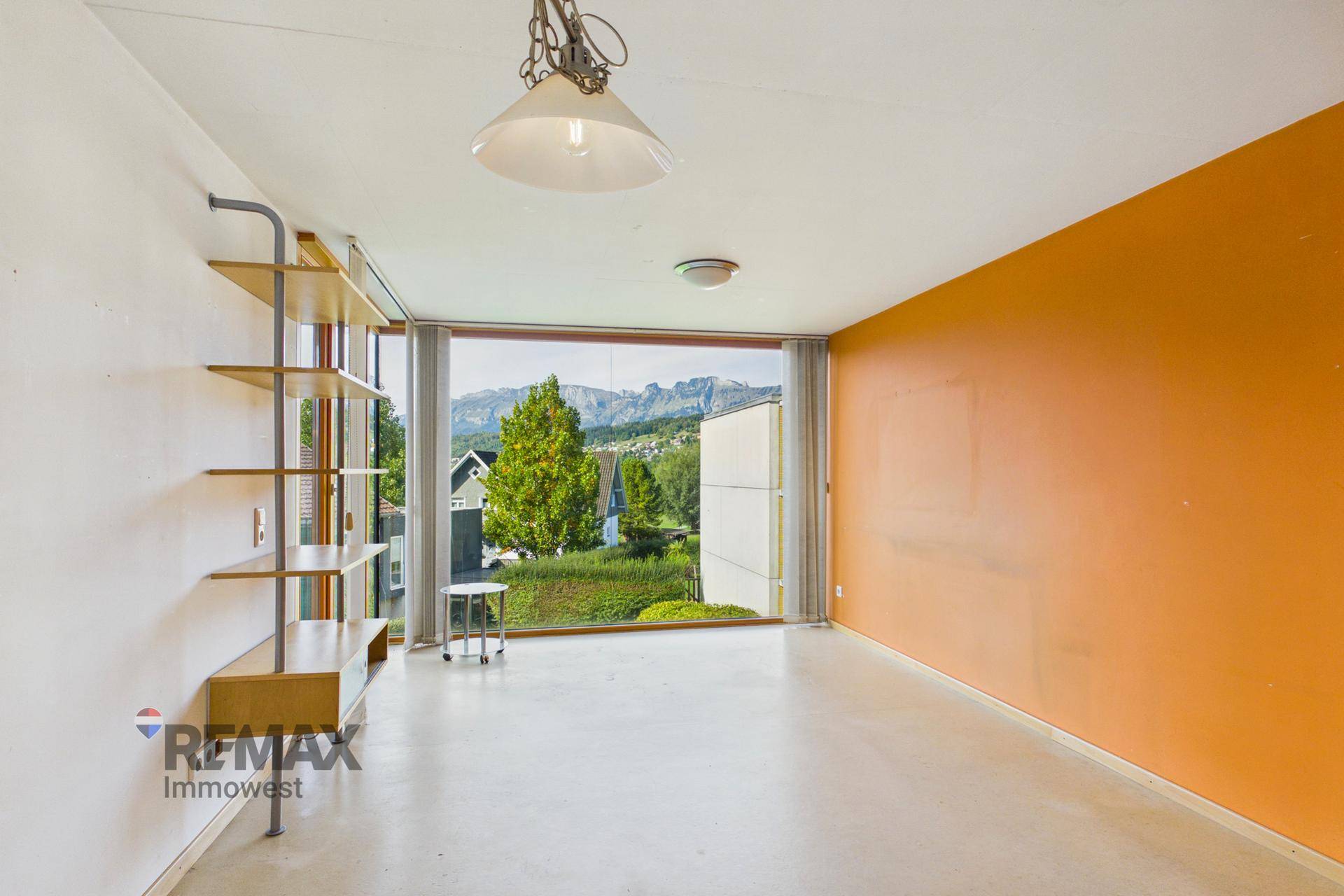 Geräumiger, leerer Wohnraum mit orangefarbener Akzentwand und weitem Bergblick.