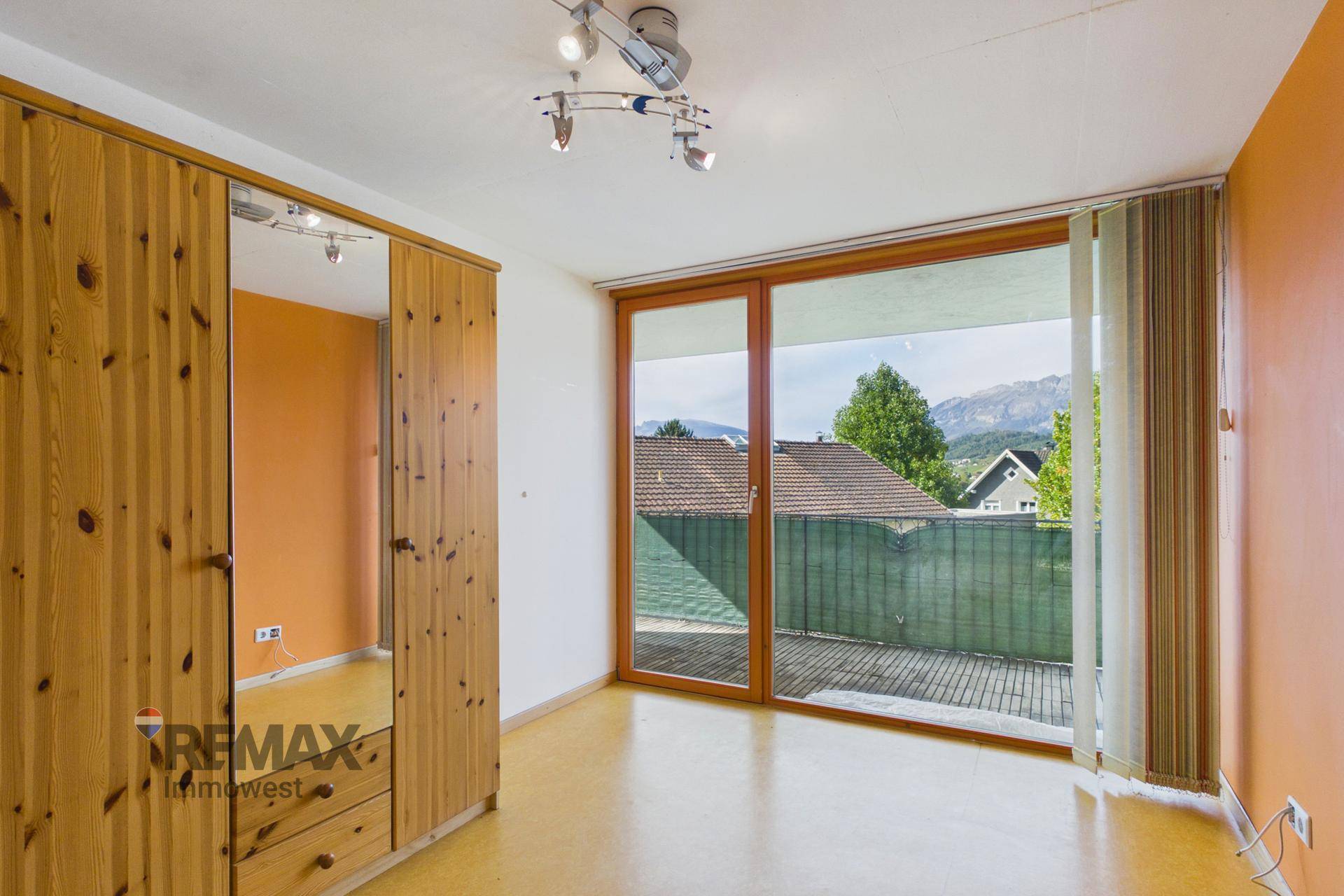 Schlafzimmer mit Holzschrank, orangefarbener Wand und Zugang zum Balkon mit Bergblick.