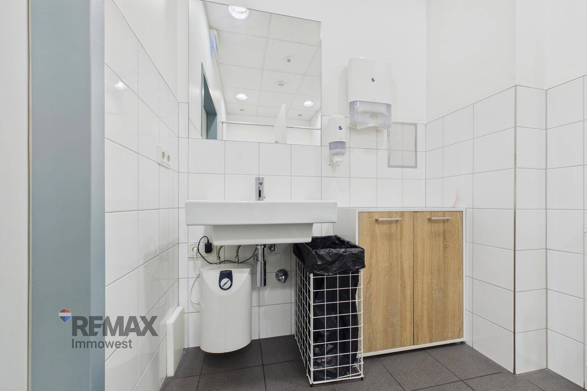 Funktionales Badezimmer mit weißen Fliesen, Waschbecken, Spiegel und einem kleinen Holzschrank.