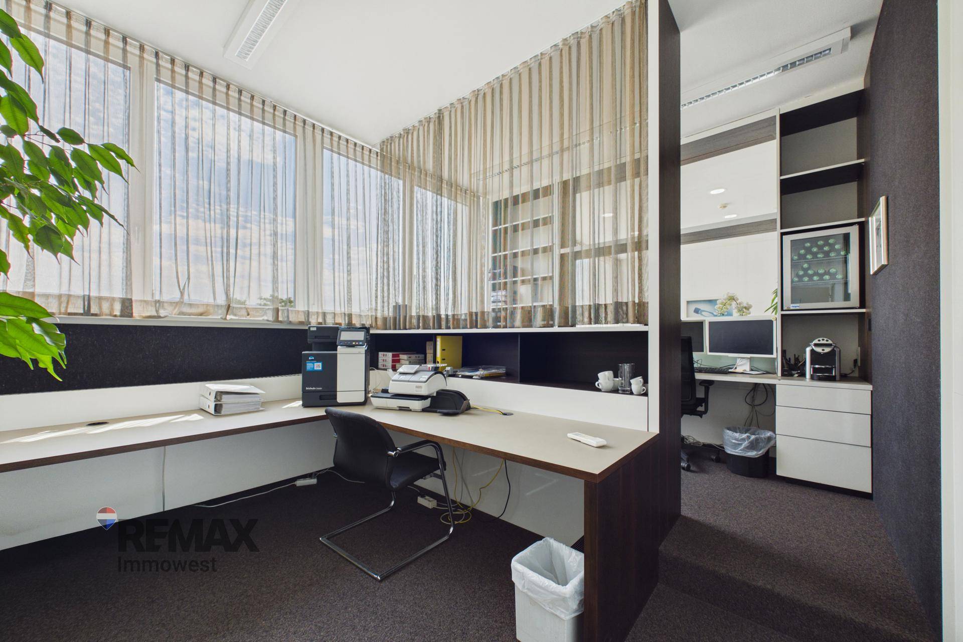 Helles Büro mit Schreibtisch, Drucker, integrierten Regalen und großen Fenstern mit Jalousien.