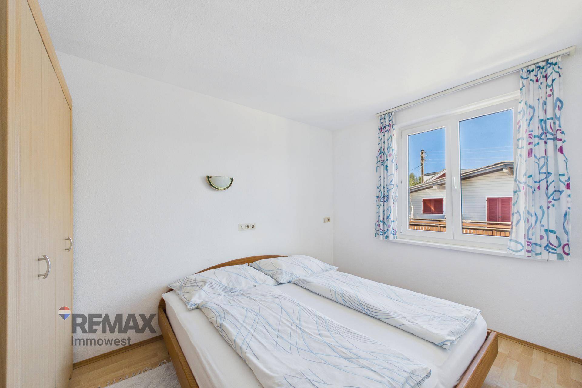 Schlafzimmer mit Doppelbett, hellem Holzschrank und Fenster mit Blick nach draußen.