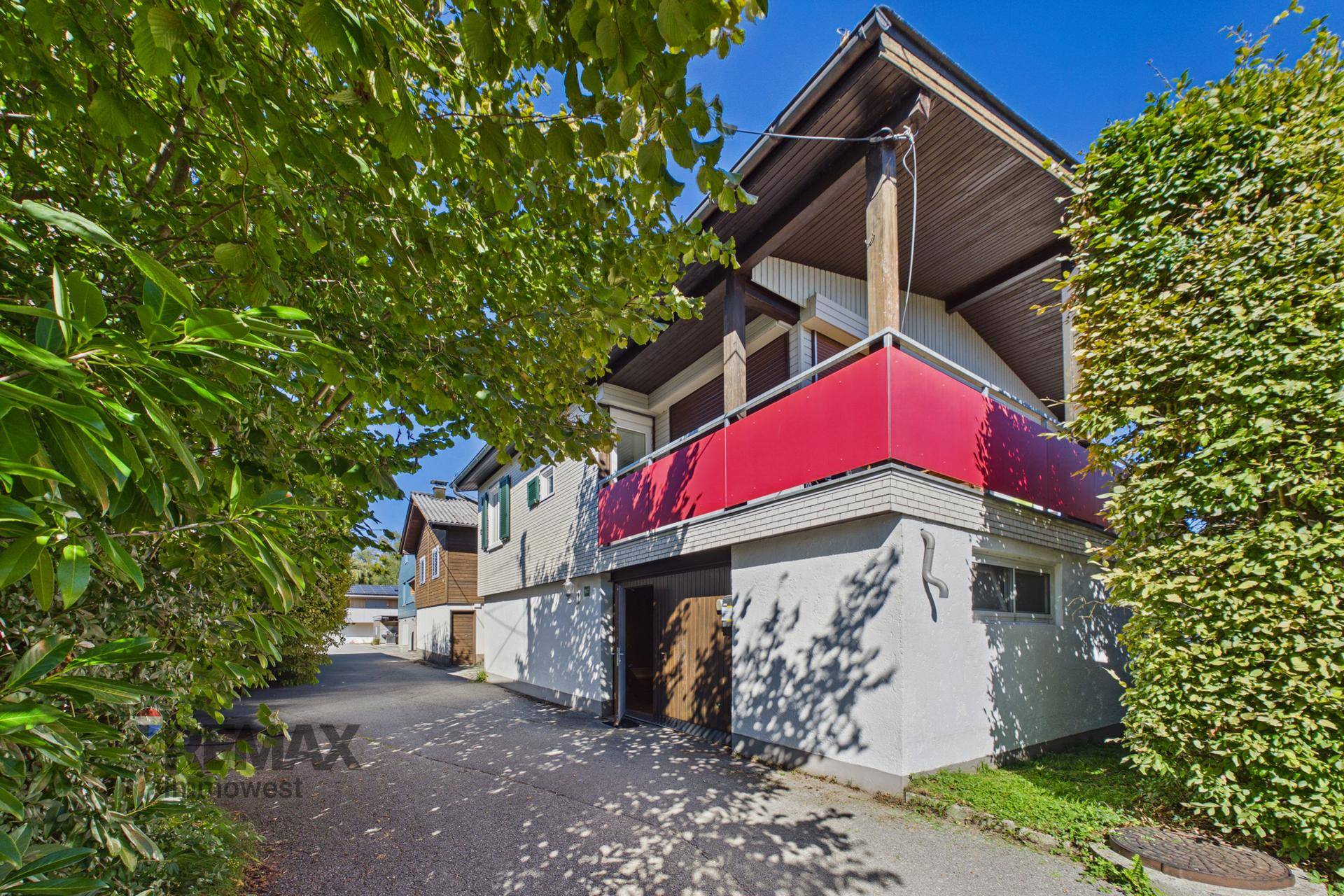 Außenansicht eines Hauses mit rotem Balkon, Zufahrt und üppiger grüner Bepflanzung.