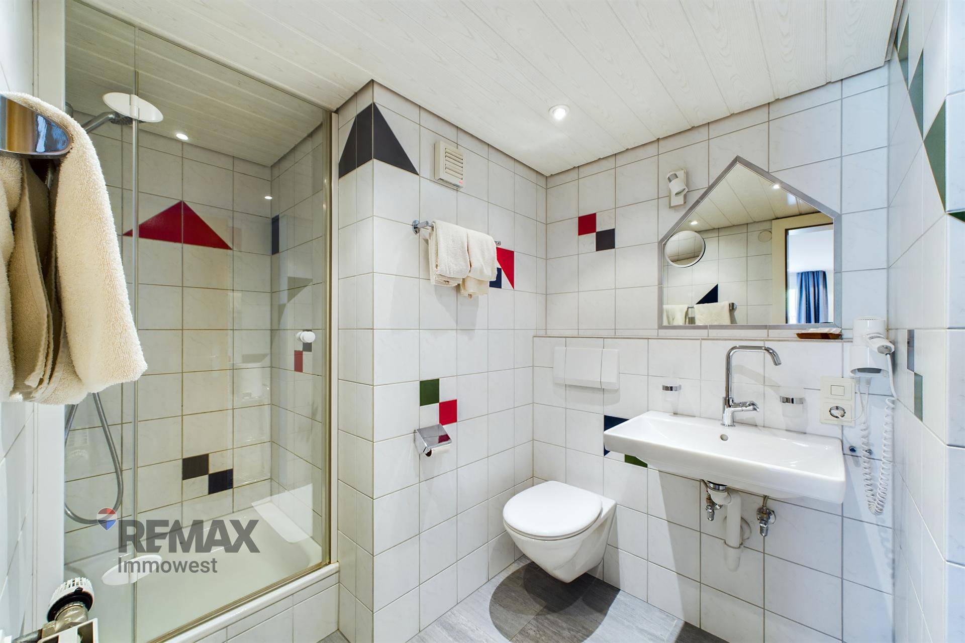 Badezimmer mit Duschkabine, WC, Waschbecken und weißen Fliesen mit farbigen Akzenten.